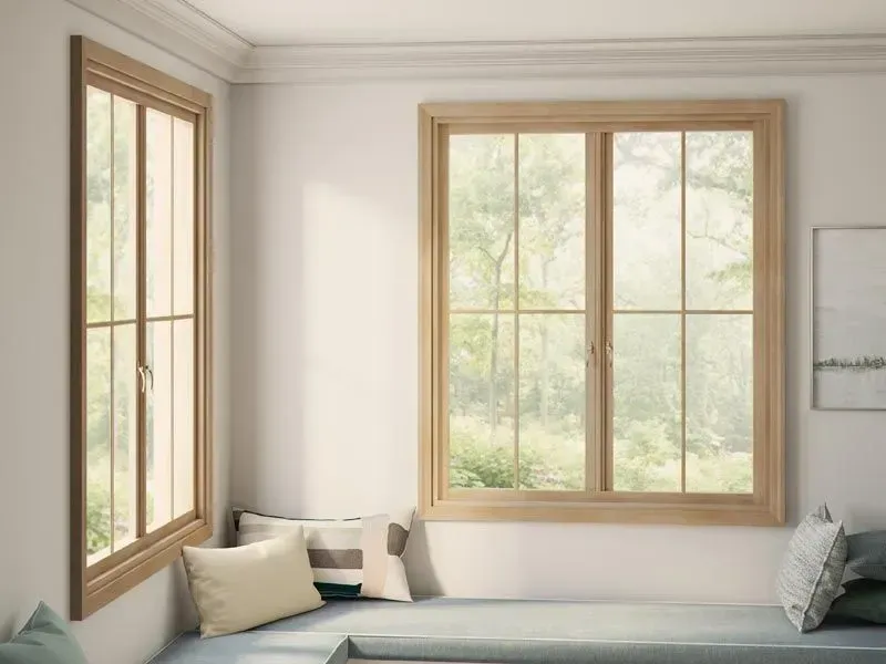 Cozy window seat in a bright room with wooden-framed windows overlooking greenery; pillows on the seat.