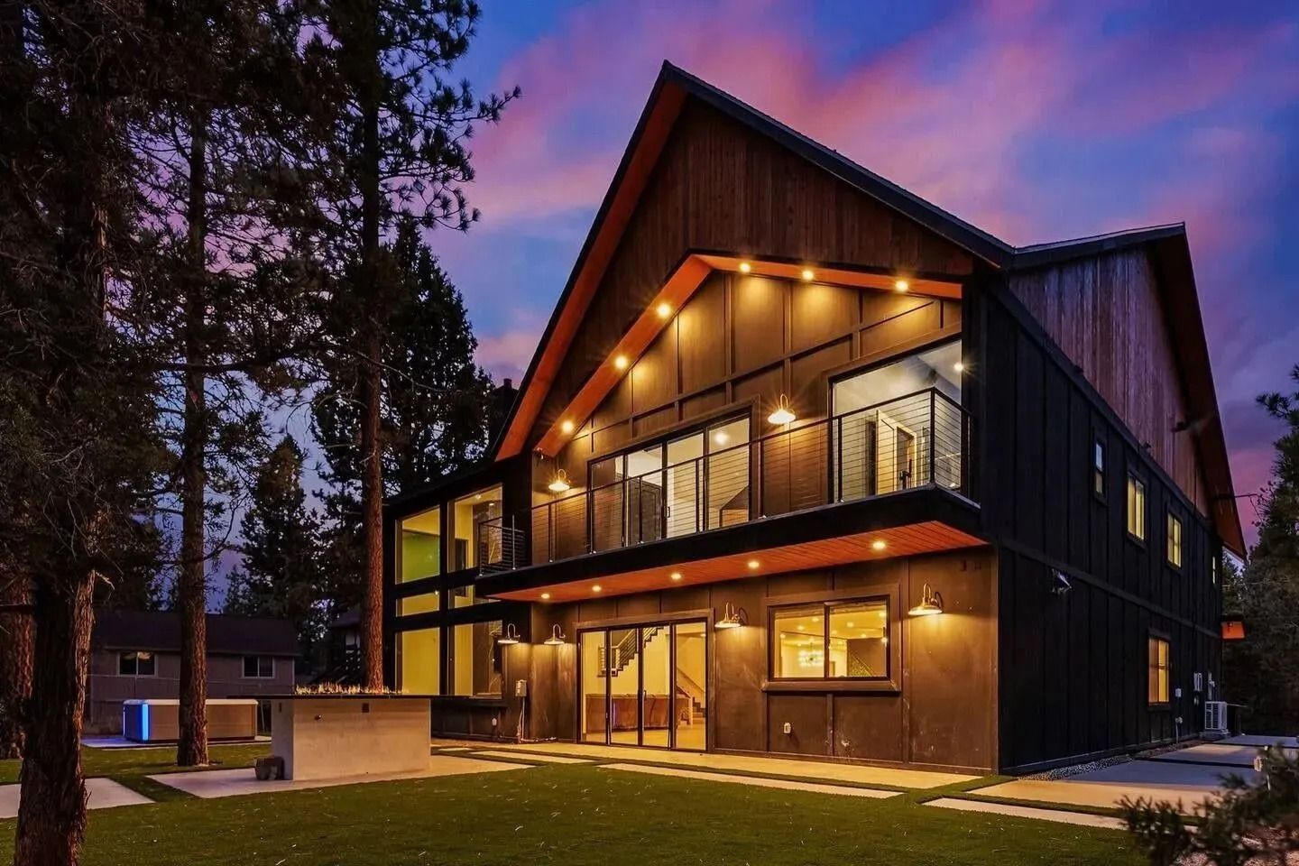 Modern two-story cabin with warm lights at dusk, large windows, and a dark wood exterior among trees.