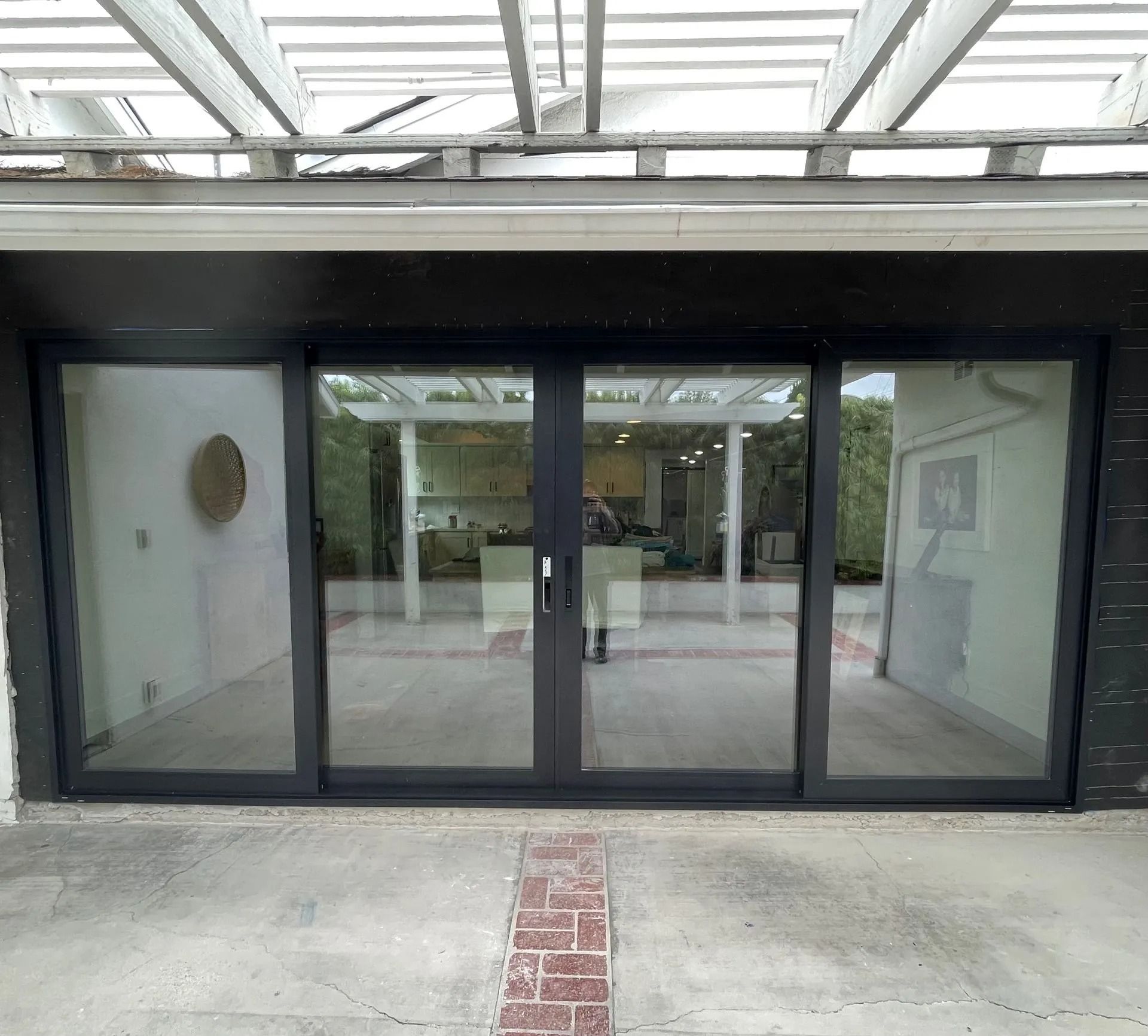 Black framed sliding glass doors on a patio, reflecting a room with a white pergola and a glimpse of the outdoors.
