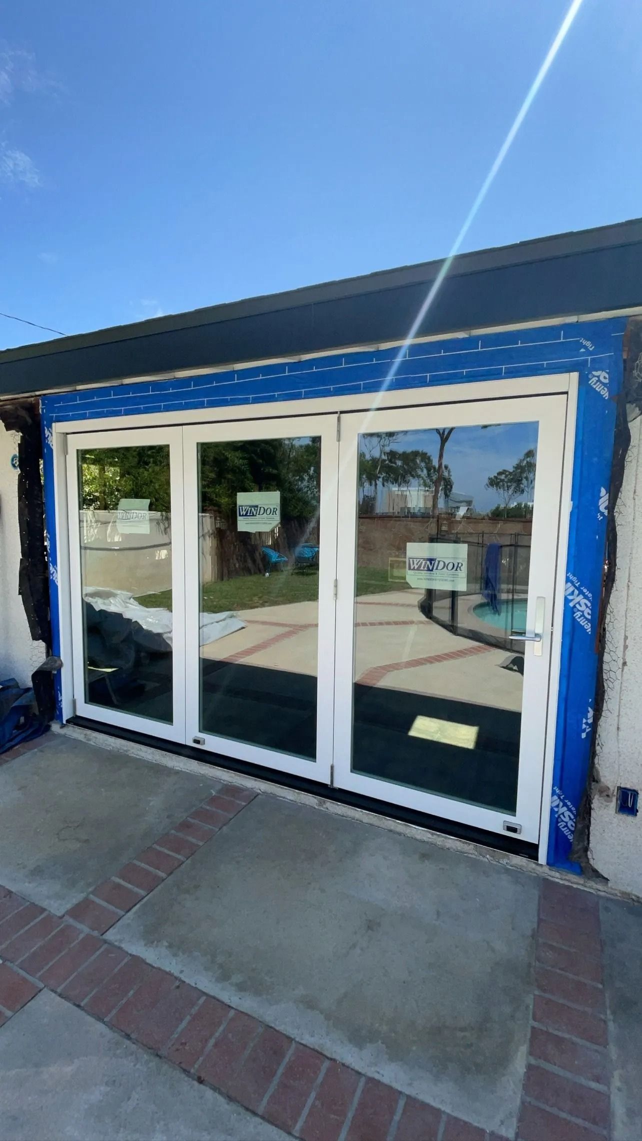 Three-panel white framed glass doors, blue tape around the edges, facing a brick patio, bright sunlight.
