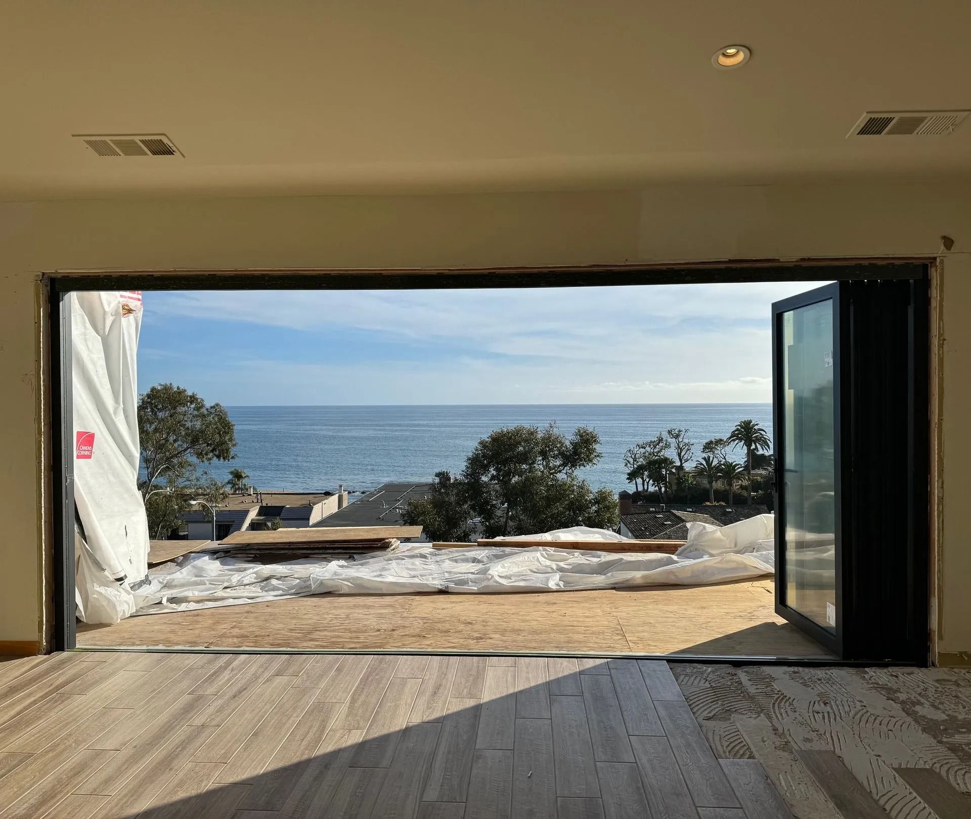 Open folding doors reveal ocean view from a room under construction; wood floor, bright sky.