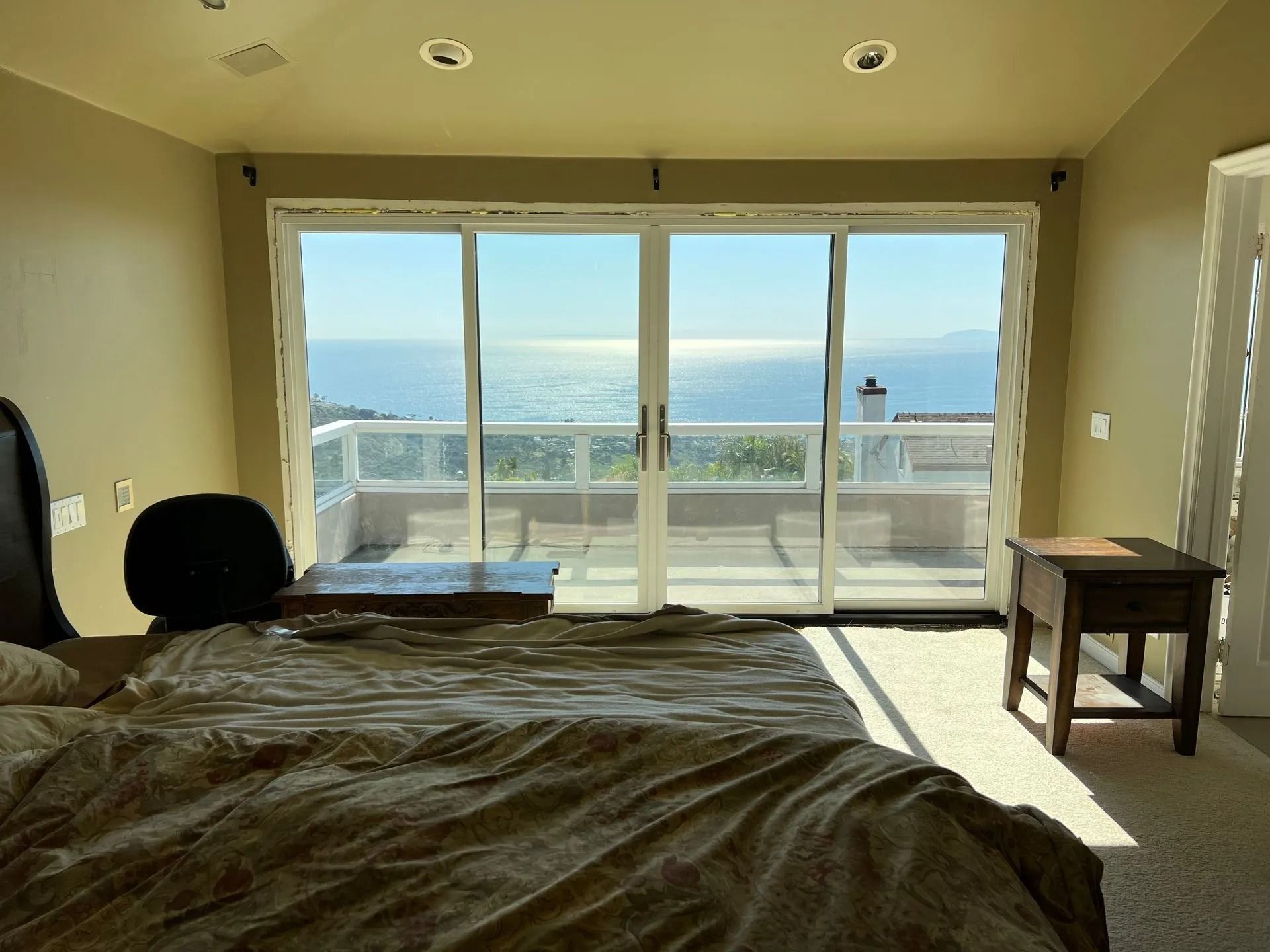 Bedroom with sliding glass doors overlooking a bright ocean view. Brown bed, nightstand, and beige walls.