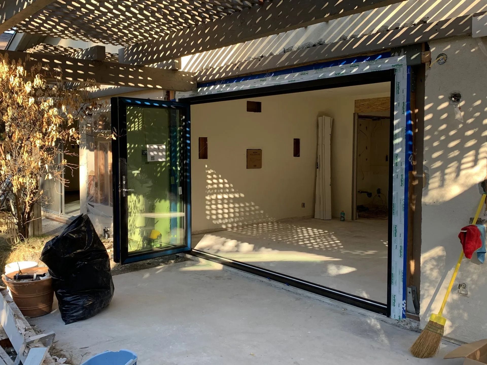 Open glass doors of a building under construction, revealing an interior. Sunlight streams through a wooden pergola.