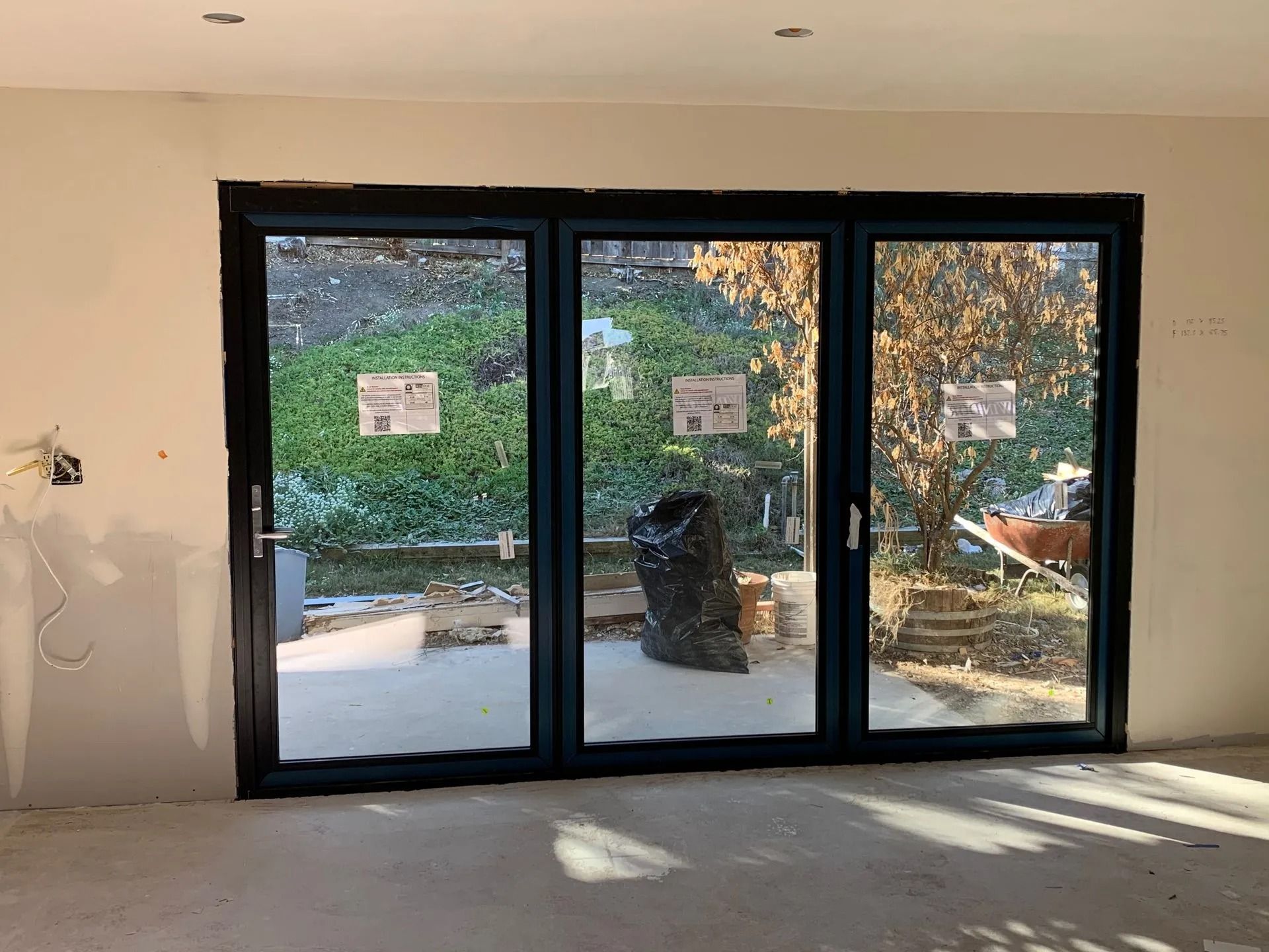 Three-panel glass door with dark frames in a room under construction, leading to a backyard.