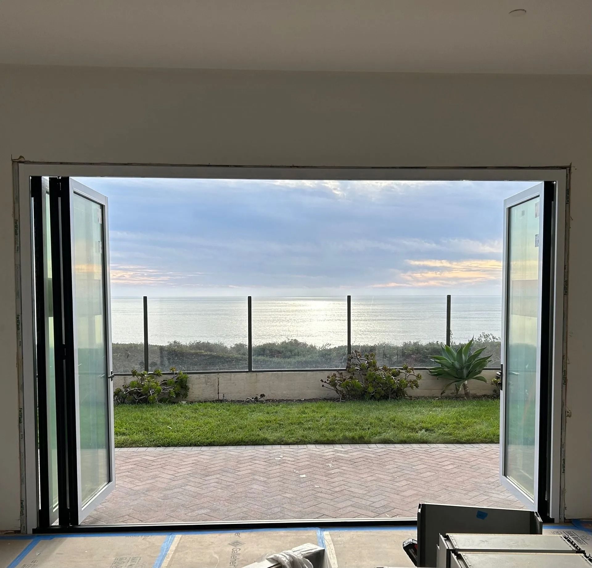 Open folding glass doors reveal a coastal view with ocean, grass, and a fence.