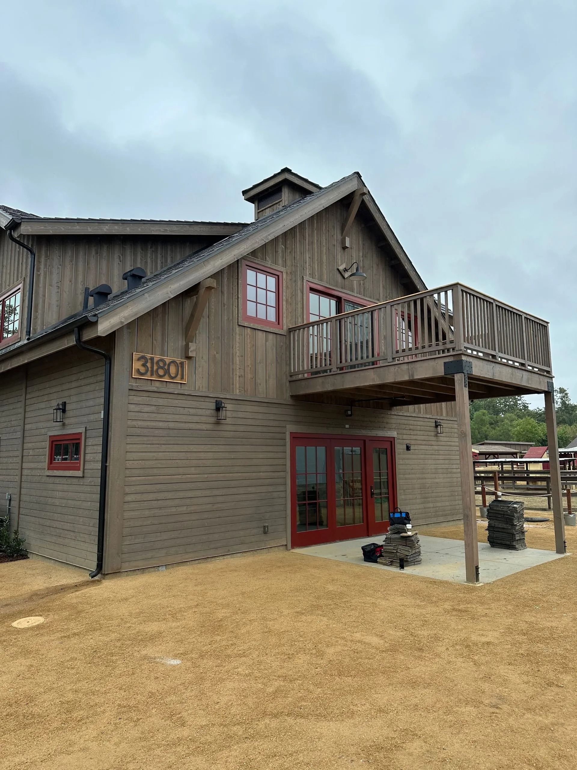 Wooden barn-style building with red-trimmed windows and a balcony; tan gravel in the foreground; cloudy sky.