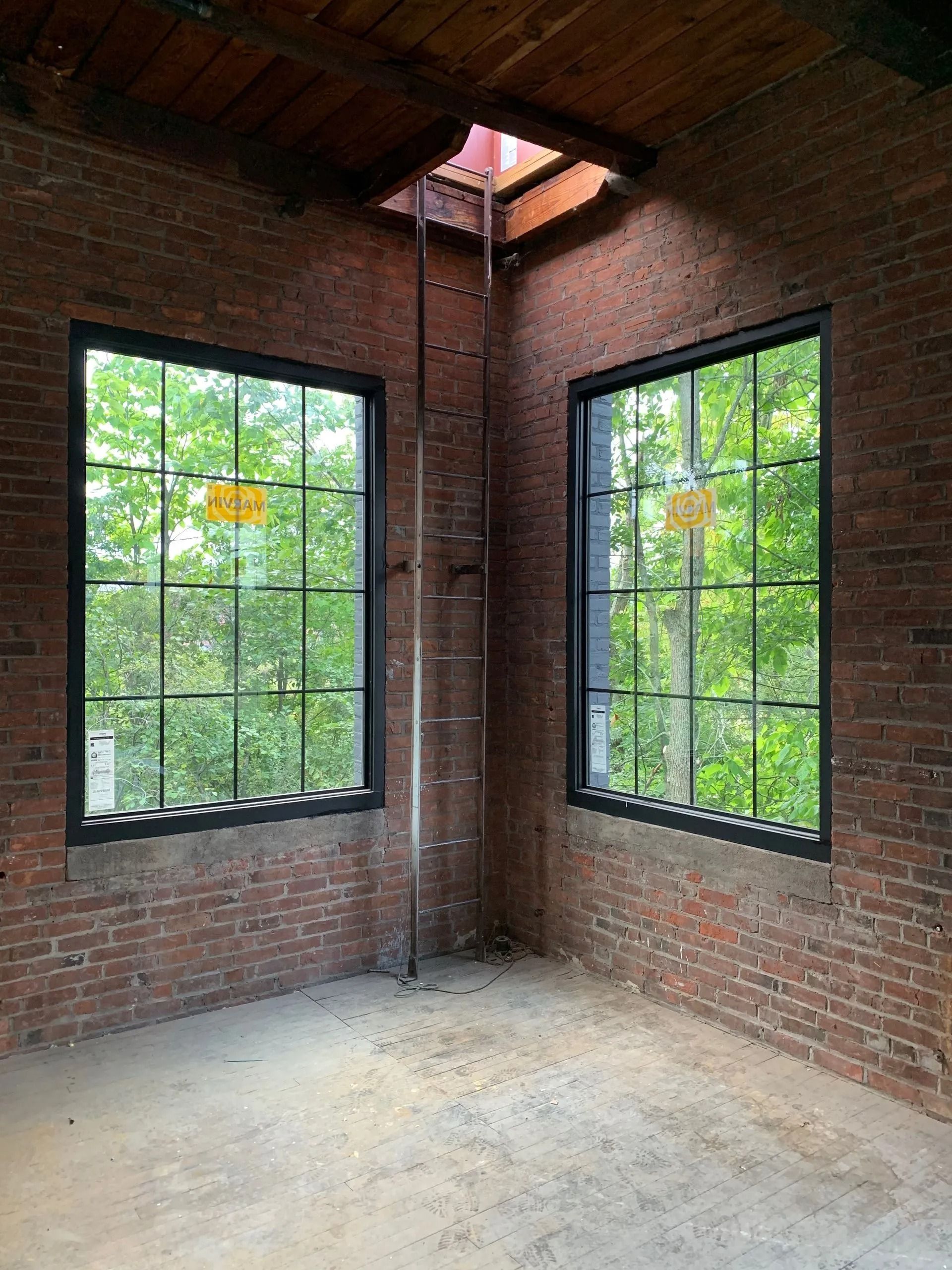 Brick-walled room with two black-framed windows, a metal ladder, and a skylight; green trees visible outside the windows.