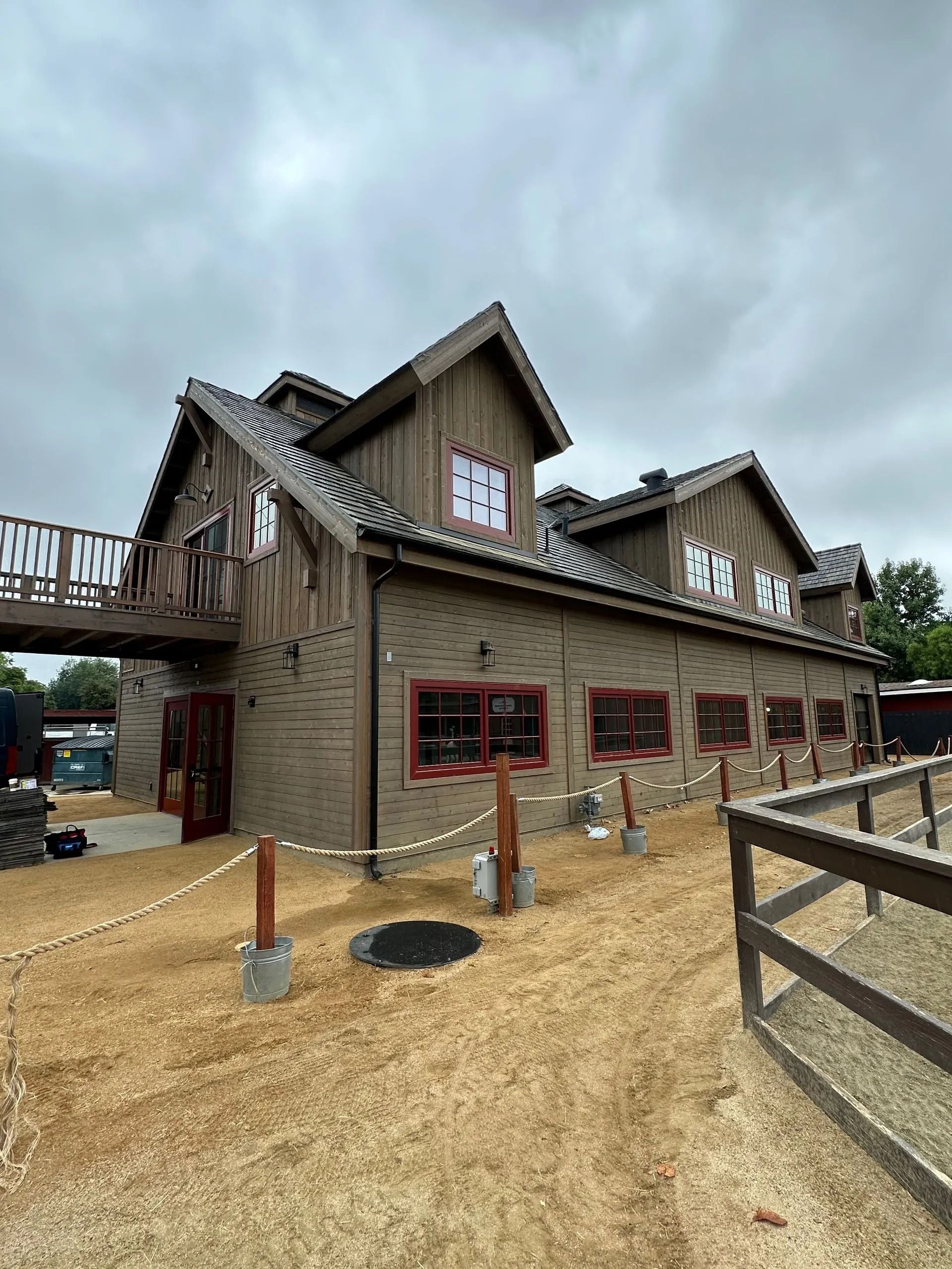 Wooden barn-style building with red-framed windows and a small balcony, on a gravel lot, with a fence.