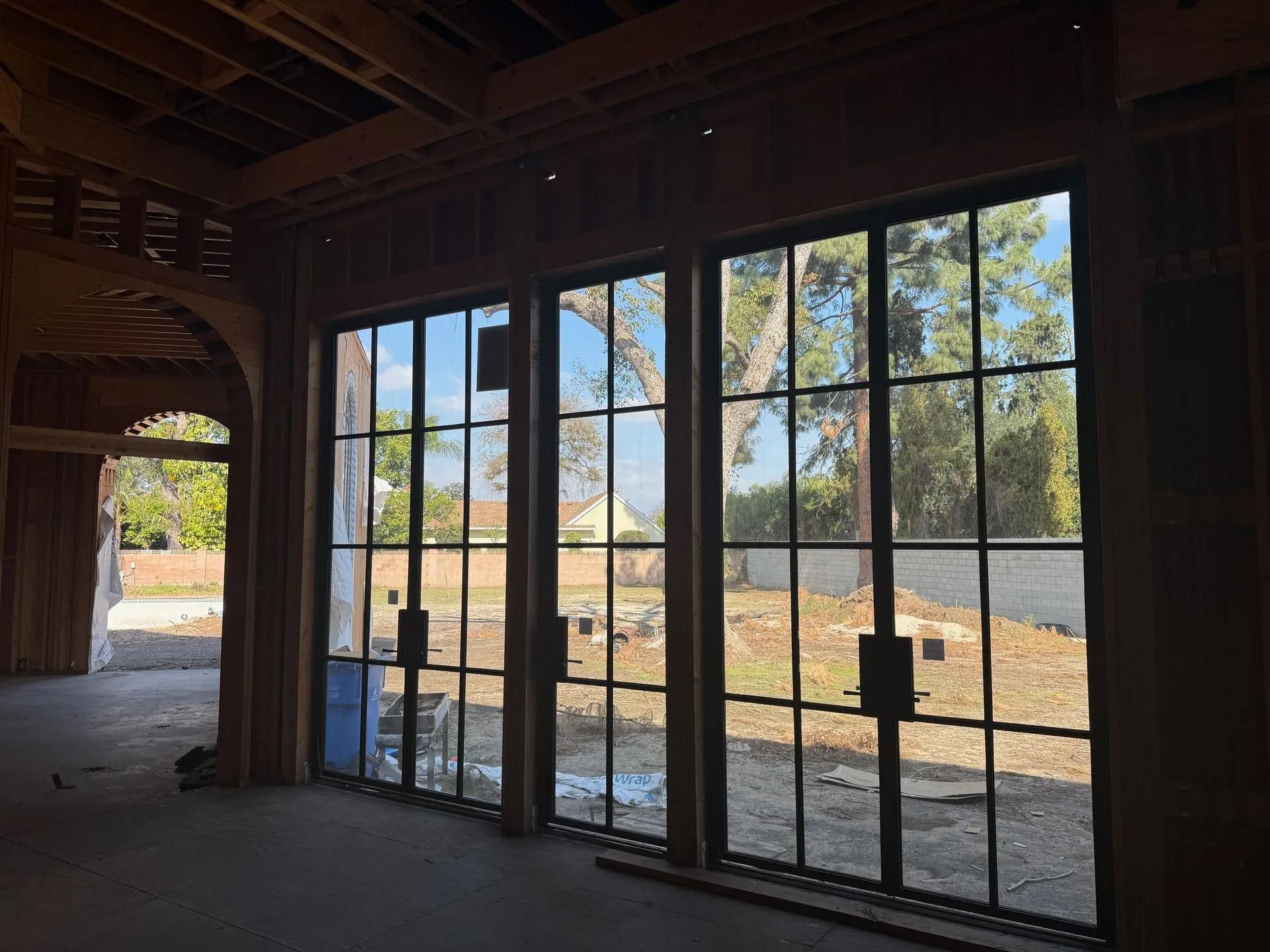 Interior view of a building under construction, featuring three large black-framed windows with a backyard view.