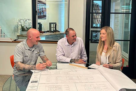 Three people review blueprints at a table. Two men and a woman look at the plans, smiling, in an office.