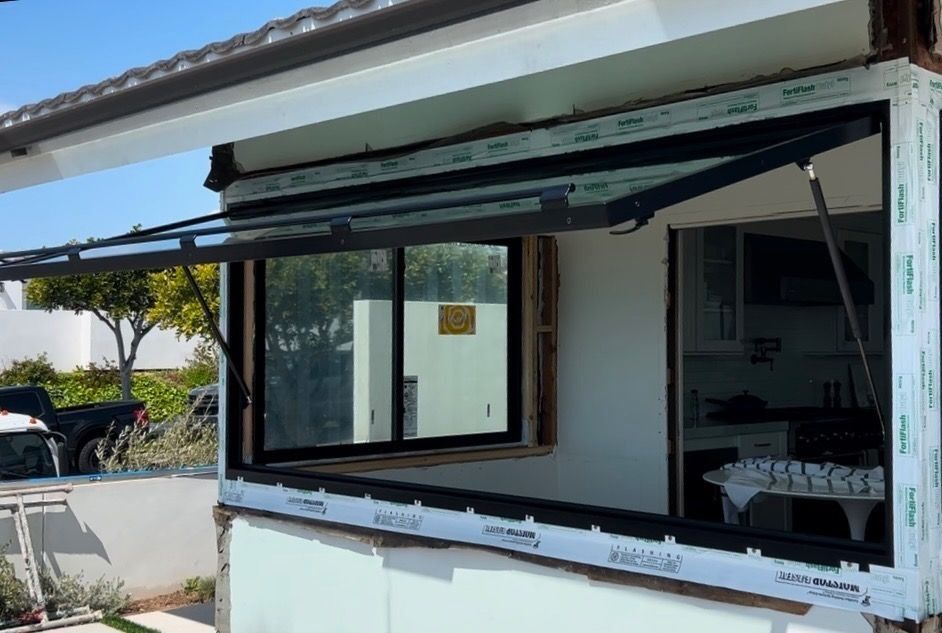 Open awning window on a white building, black frame, hinges, and supports. Green lawn and sunny outdoor setting.