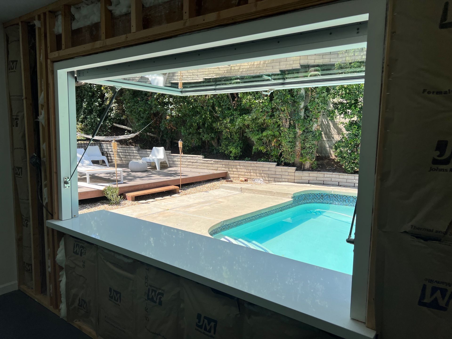 Construction view: open window frame revealing a pool, patio, and lush greenery. White trim, blue water.