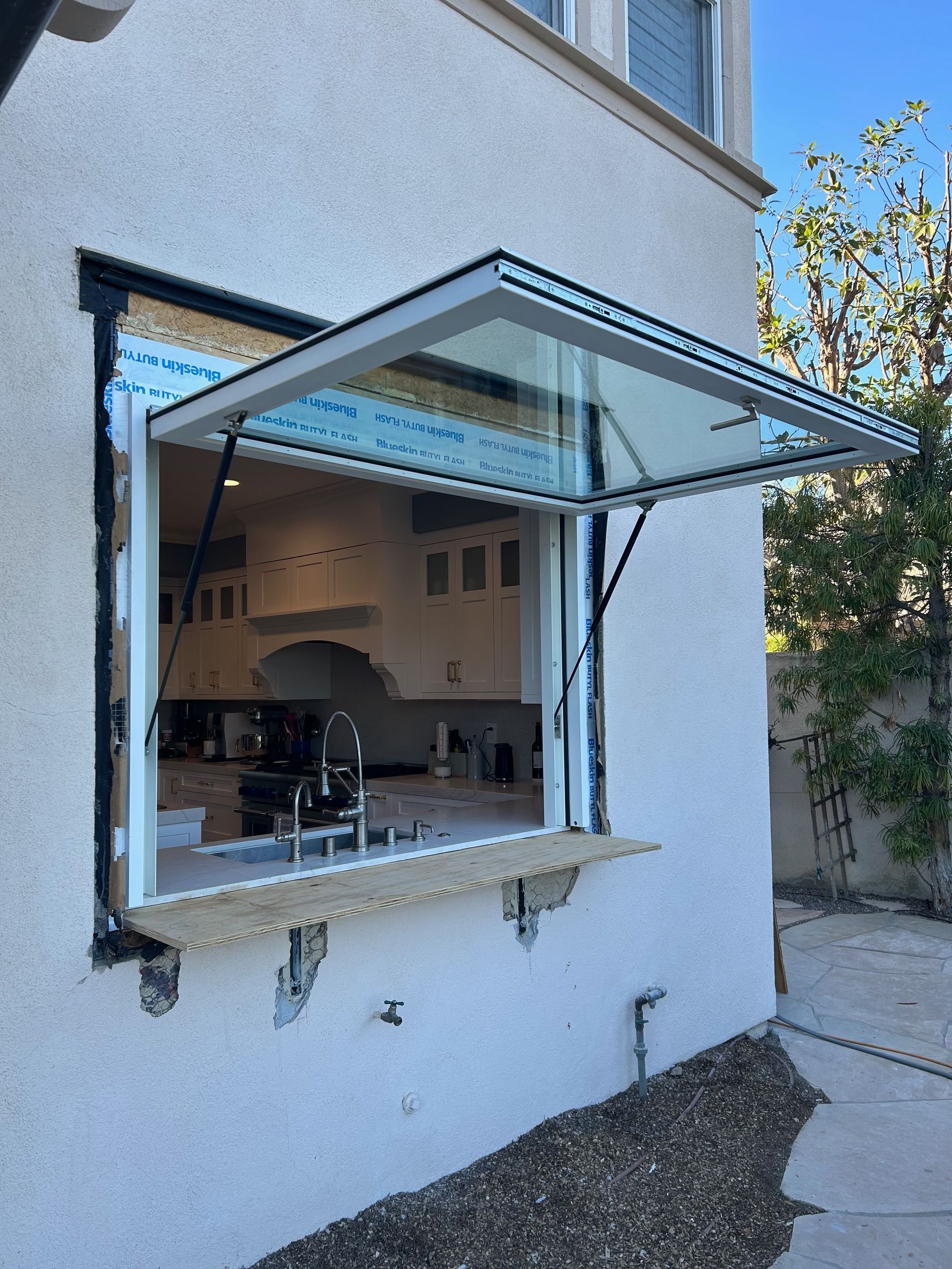 Exterior window with a glass pane that opens upward, next to a kitchen. Beige stucco wall, blue sky.