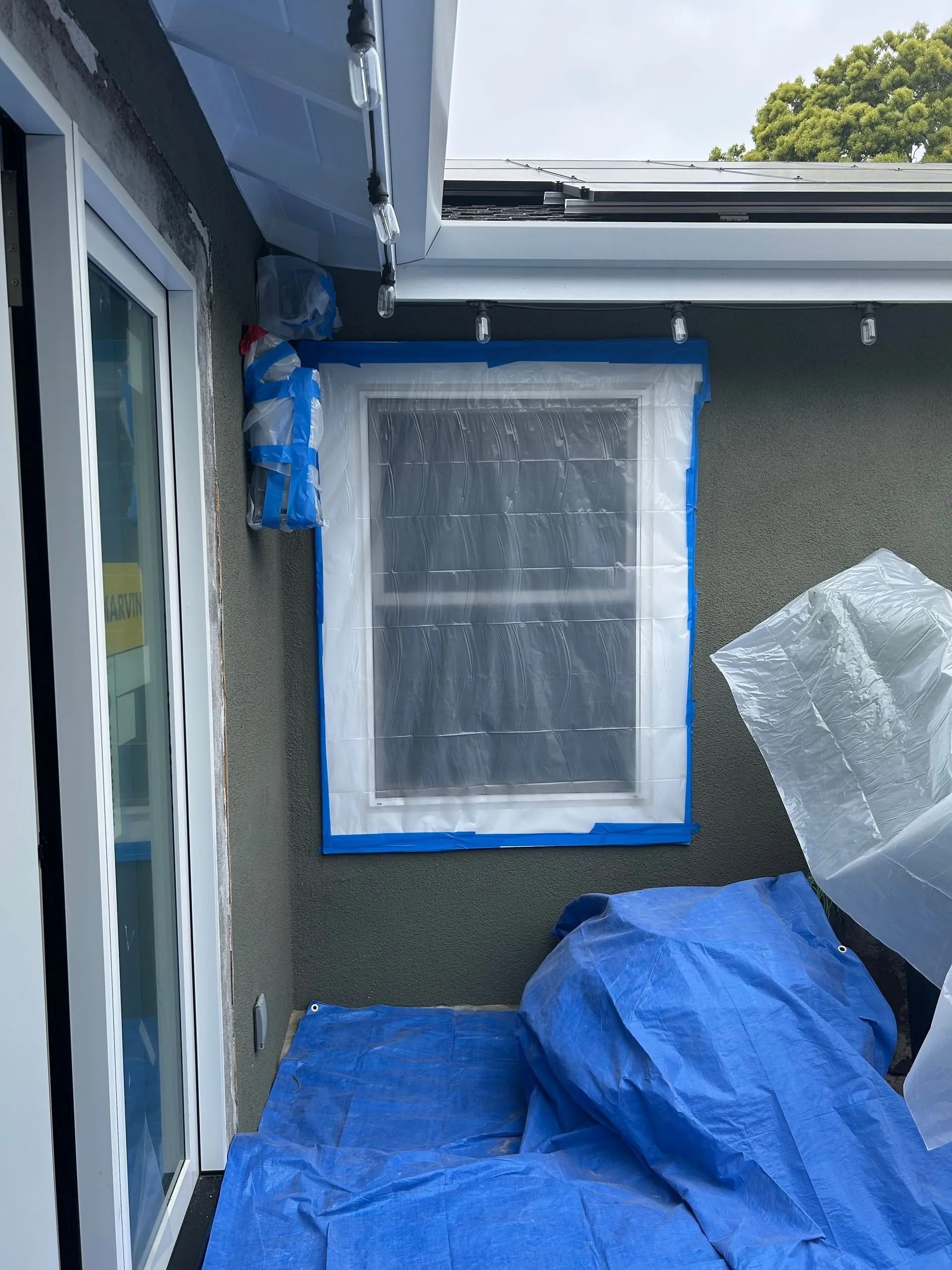 Window covered with plastic and blue tape during exterior renovation, tarp on ground.