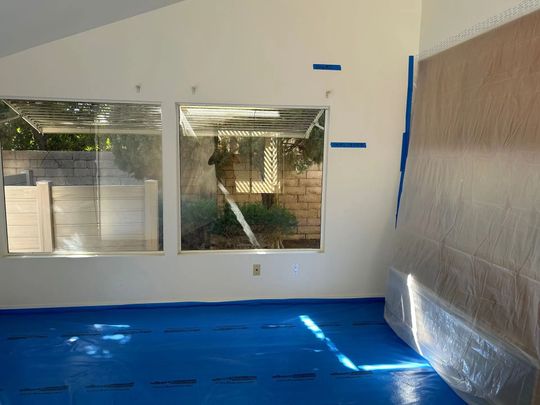 Room with blue floor covering, windows covered in plastic, wall partially covered, and tape markings.
