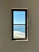 Window with black frame revealing blue sky, ocean, and white sand, set in a beige wall.