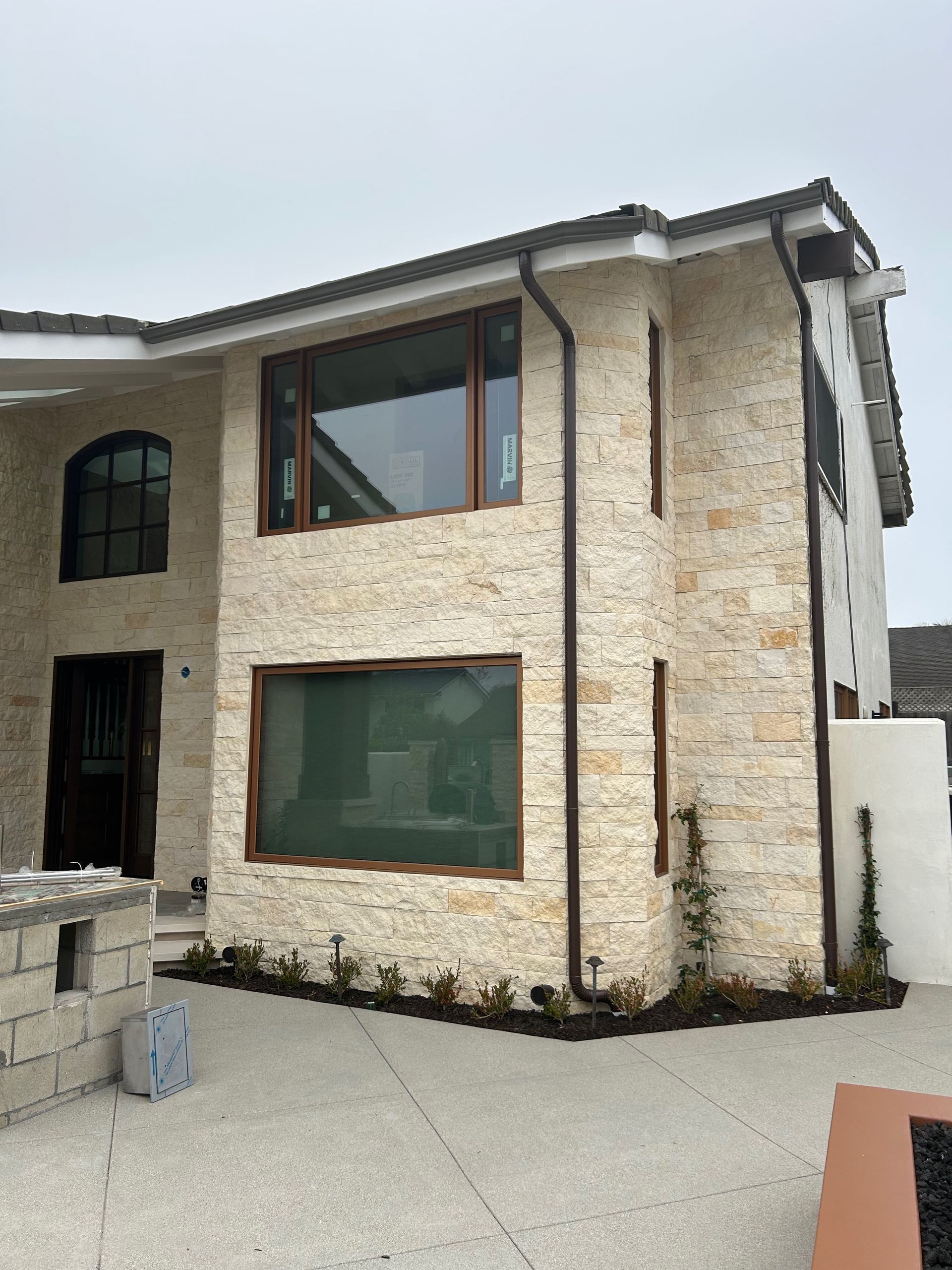 Two-story house with light stone facade, brown window frames, and paved driveway.