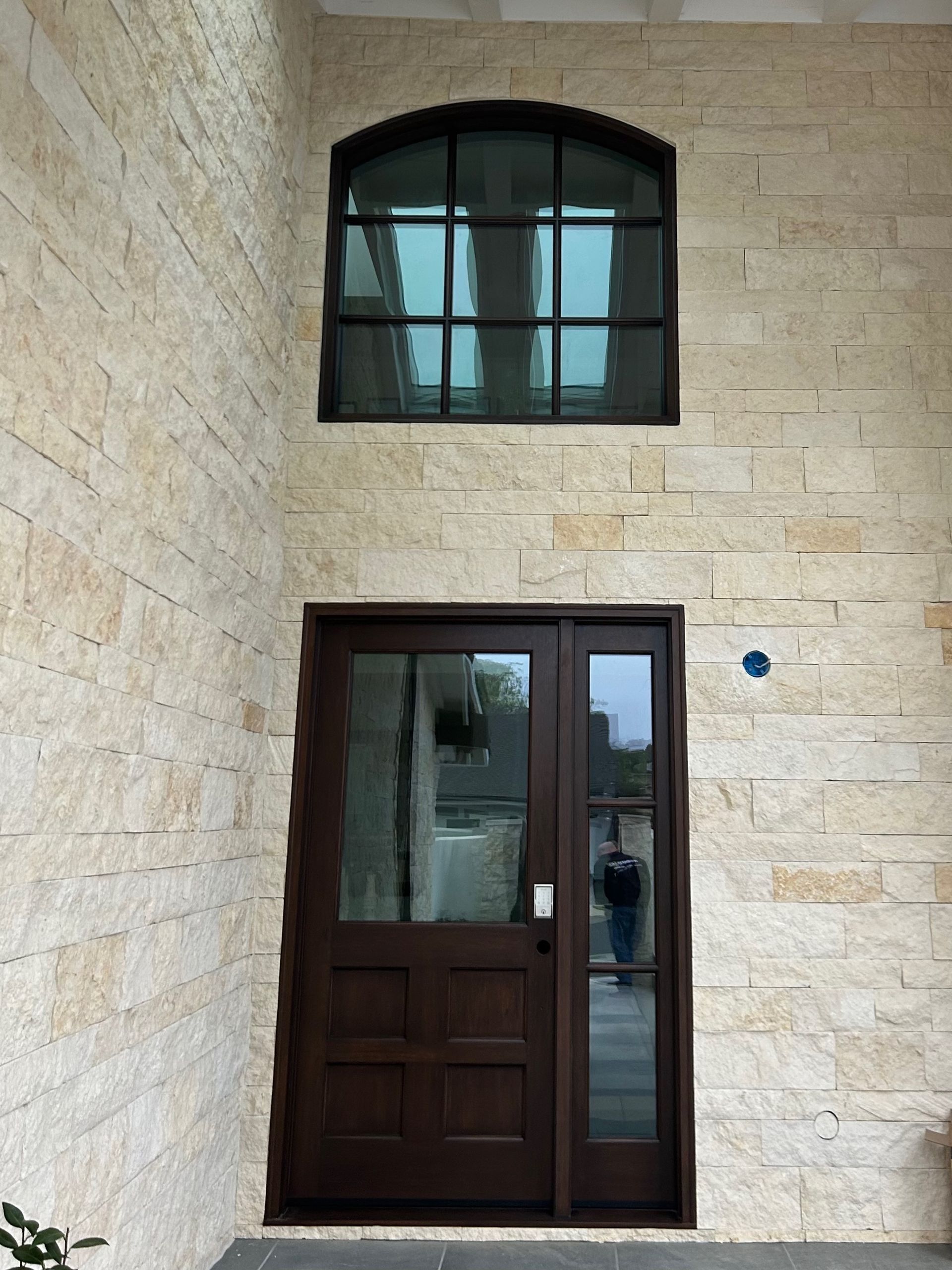 Brown front door with sidelight and arched window above, set in a light stone wall.