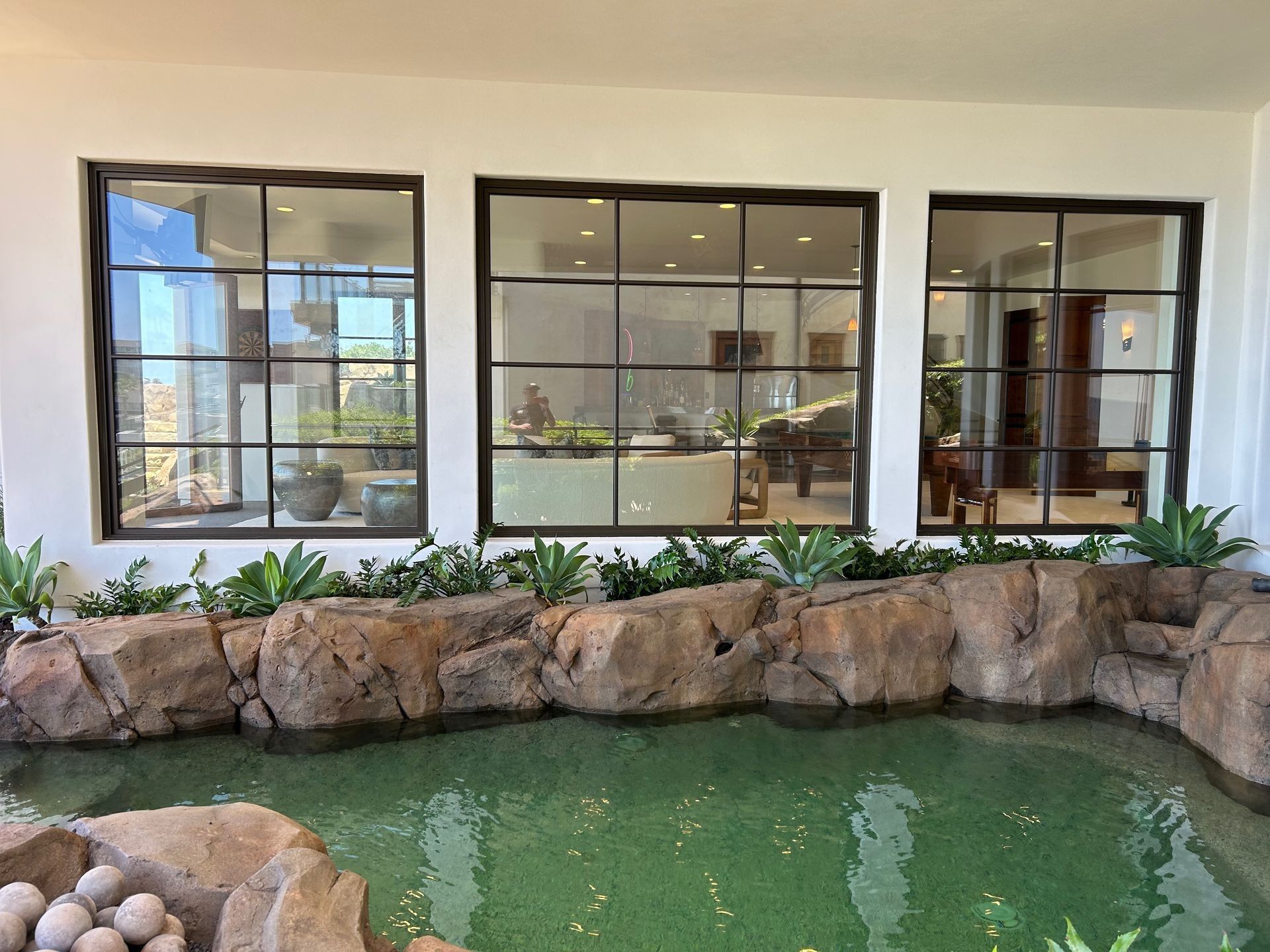 Three dark-framed windows overlook a water feature with rocks and plants. Interior shows a living space.