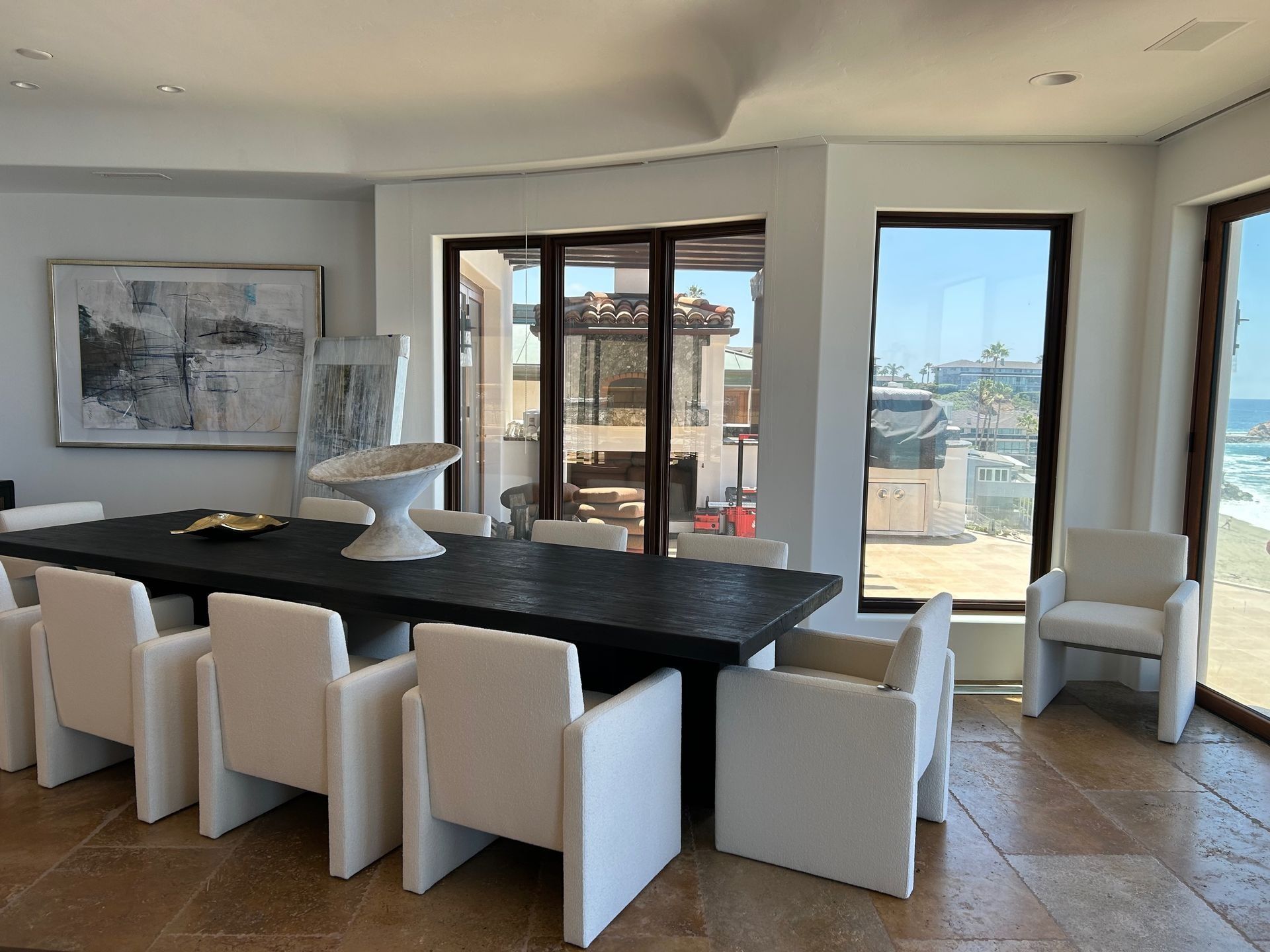Dining room with long, dark table and white chairs; ocean view visible through windows.