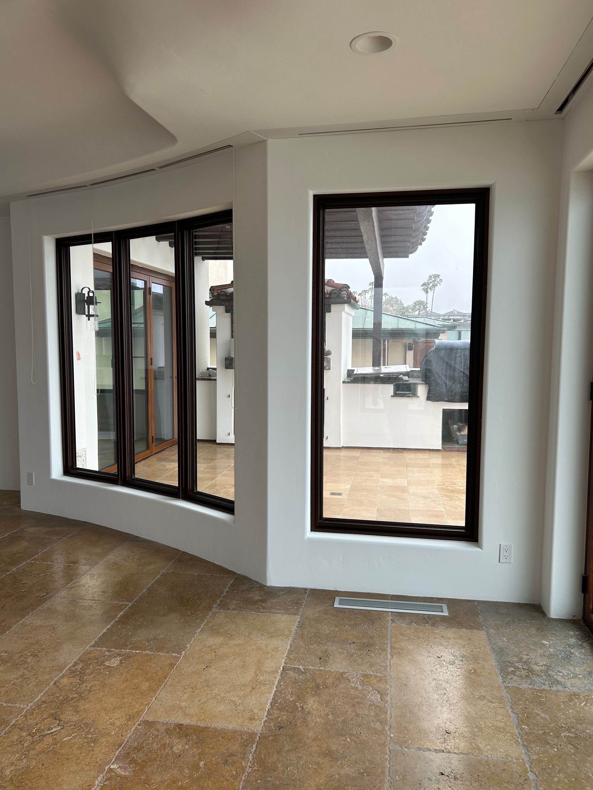 Interior room with large windows framed in dark wood, overlooking a patio. Stone flooring, white walls.