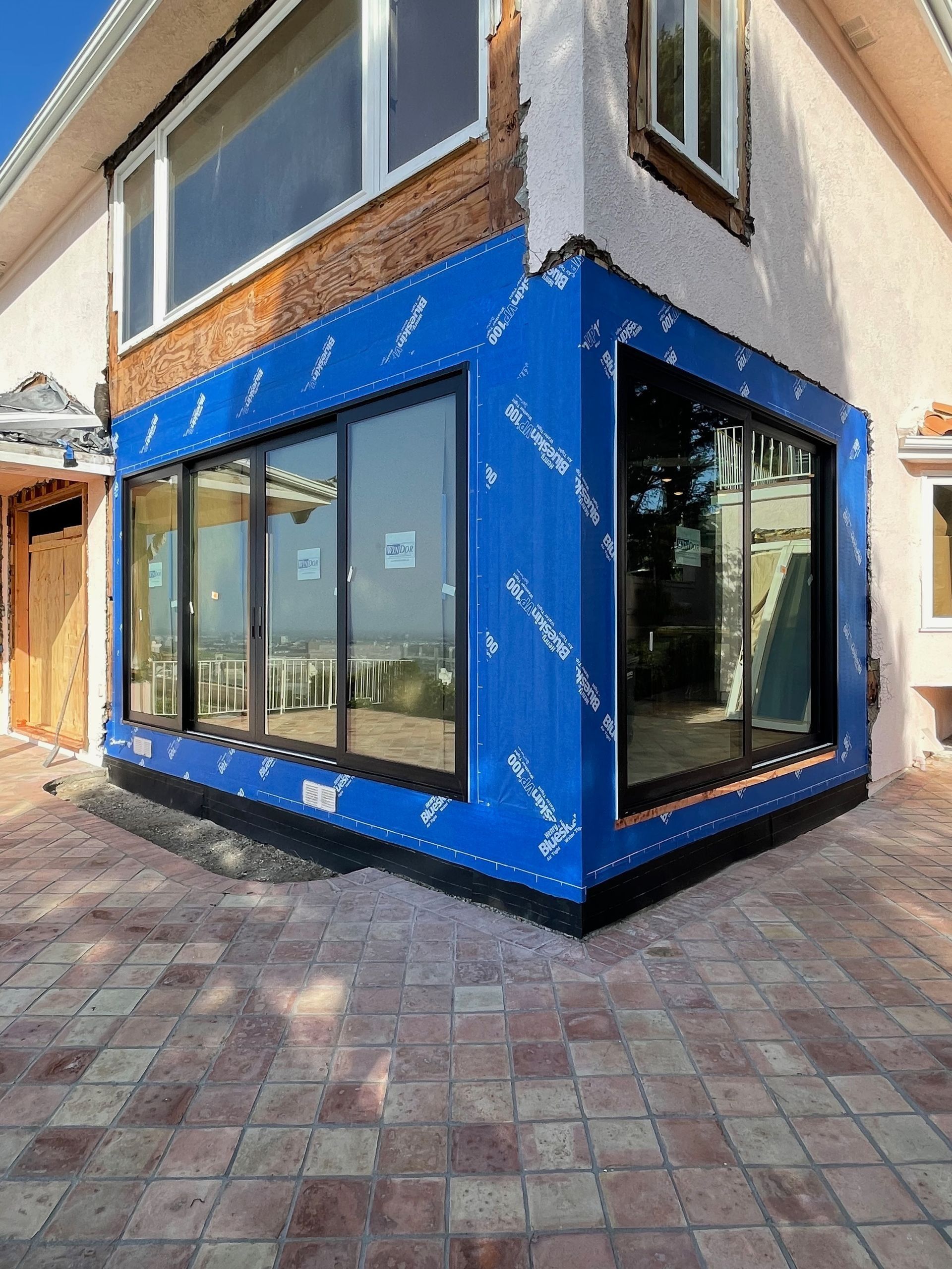 Exterior of a house under construction. Blue weather barrier wraps a corner with black framed windows. Brick and stucco walls.