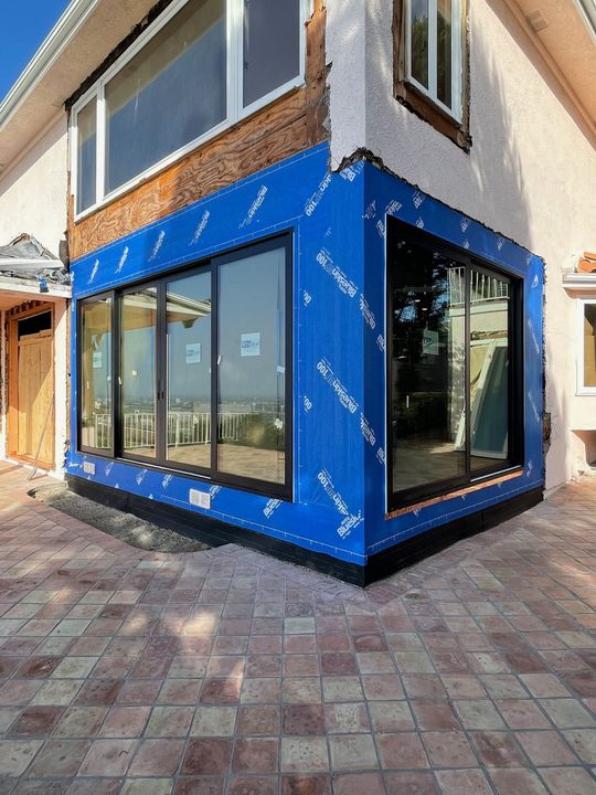 Exterior house corner with blue wrap, large black-framed windows, brick, and stucco walls, on a brick patio.