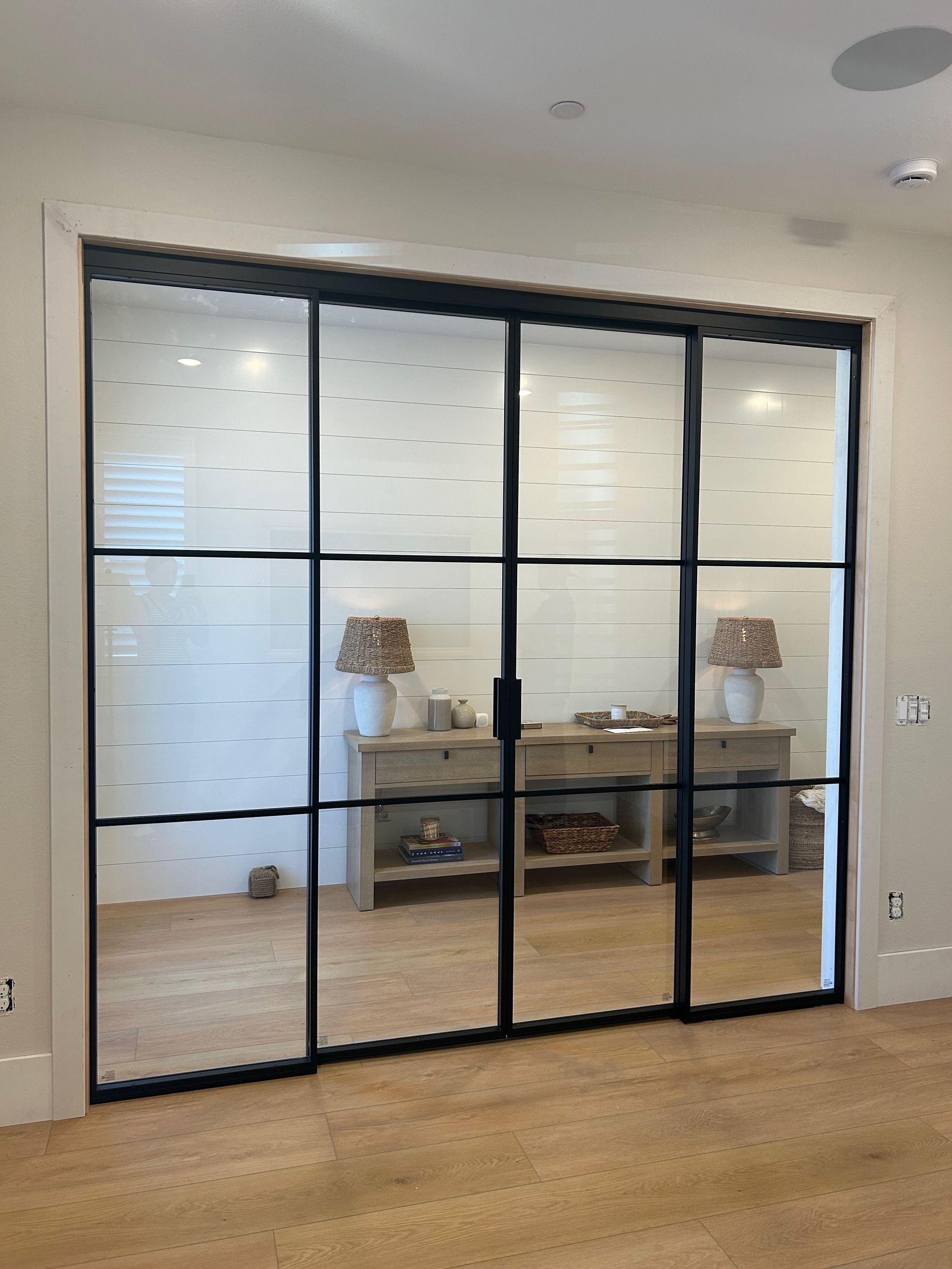 Black-framed glass sliding doors separate a room. Behind the doors is a console table with lamps.