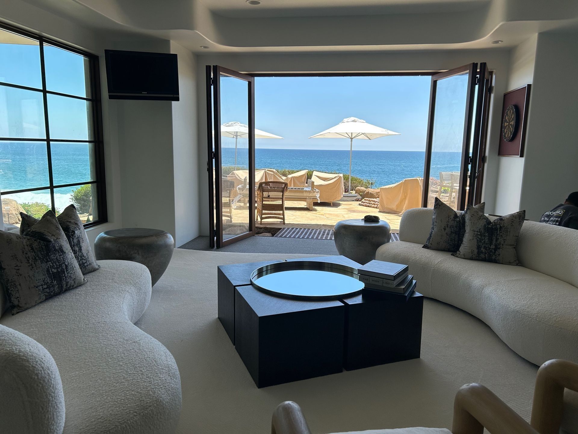 Living room with ocean view: curved white couches, black square coffee table, open doors to patio with umbrellas.