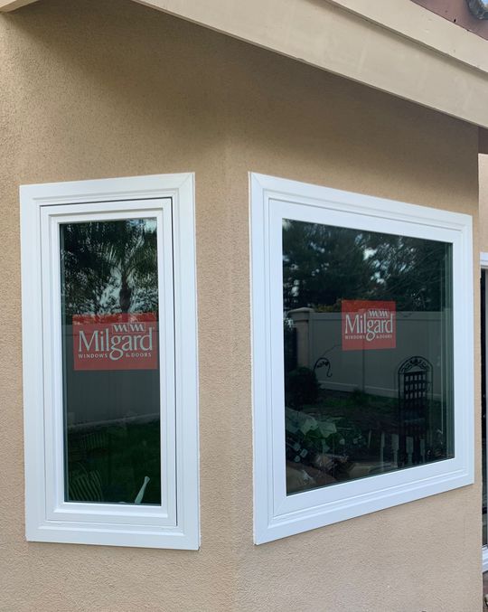 Two white-framed windows on a beige stucco wall; each window has a Milgard label inside.