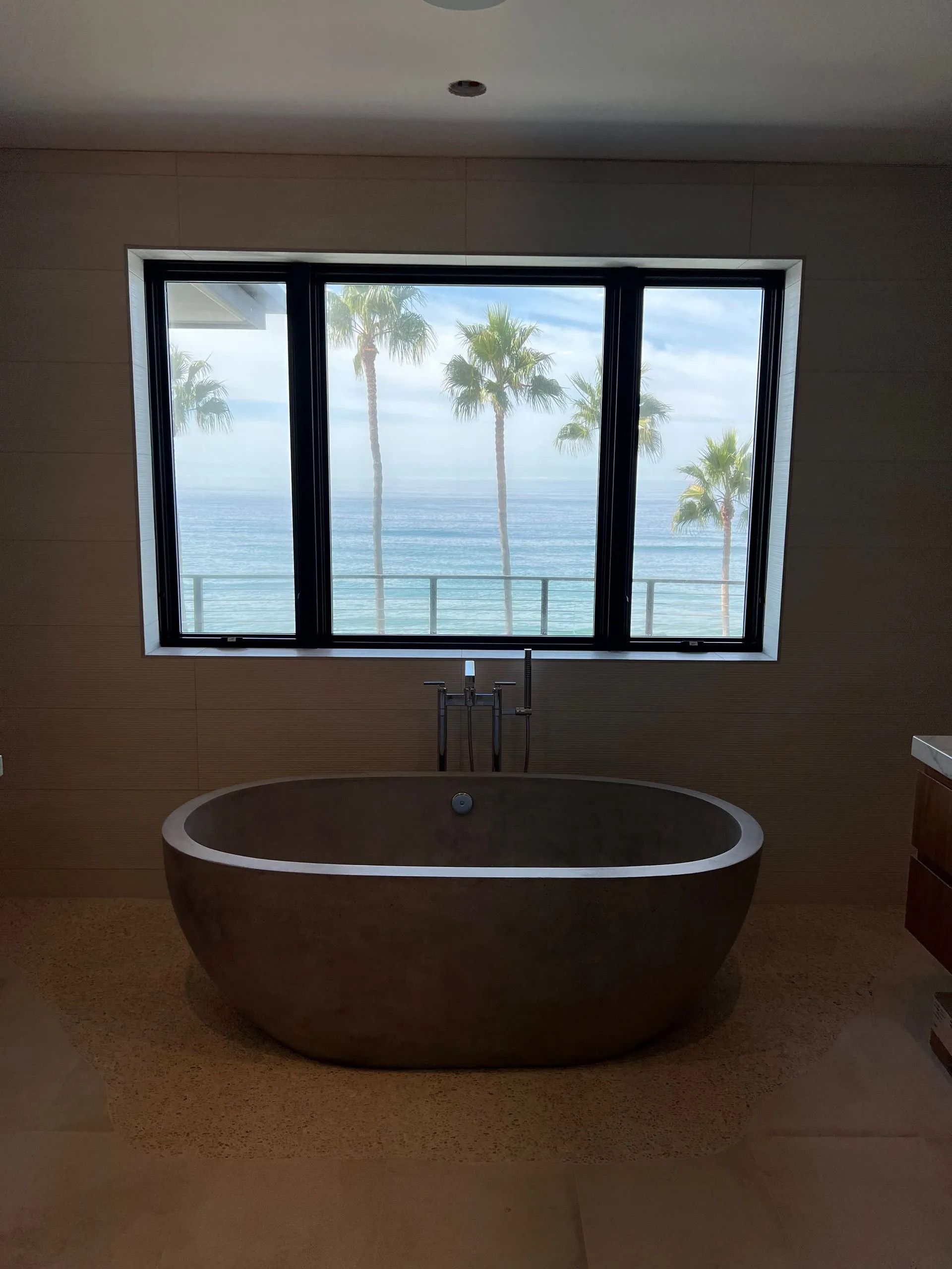 Bathroom with a dark oval tub in front of a window overlooking the ocean and palm trees.