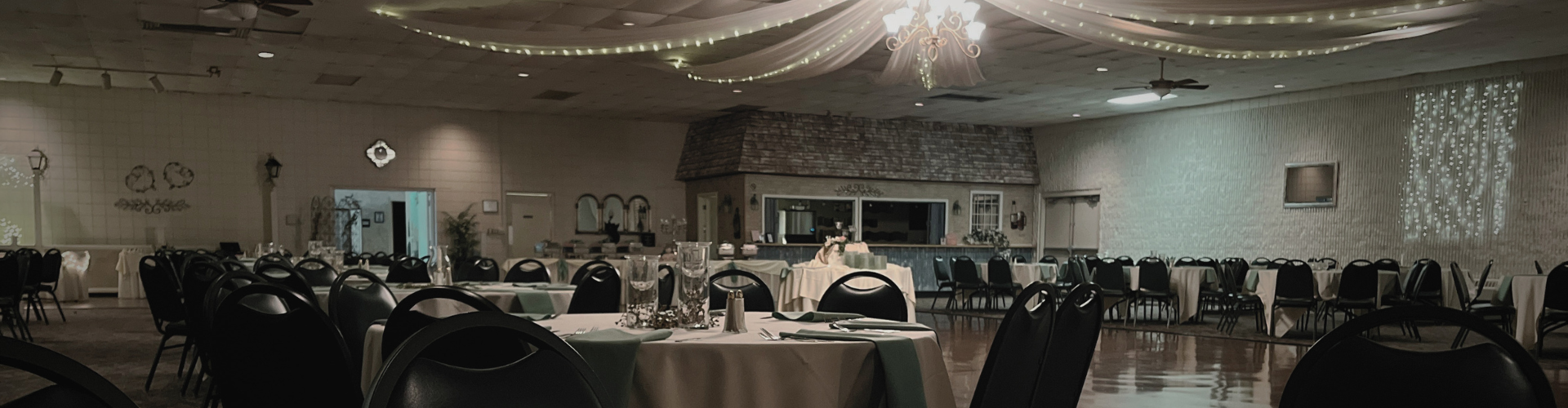 A wide-angle interior view of a banquet hall featuring round tables with white cloths, black chairs, and ceiling drapery.