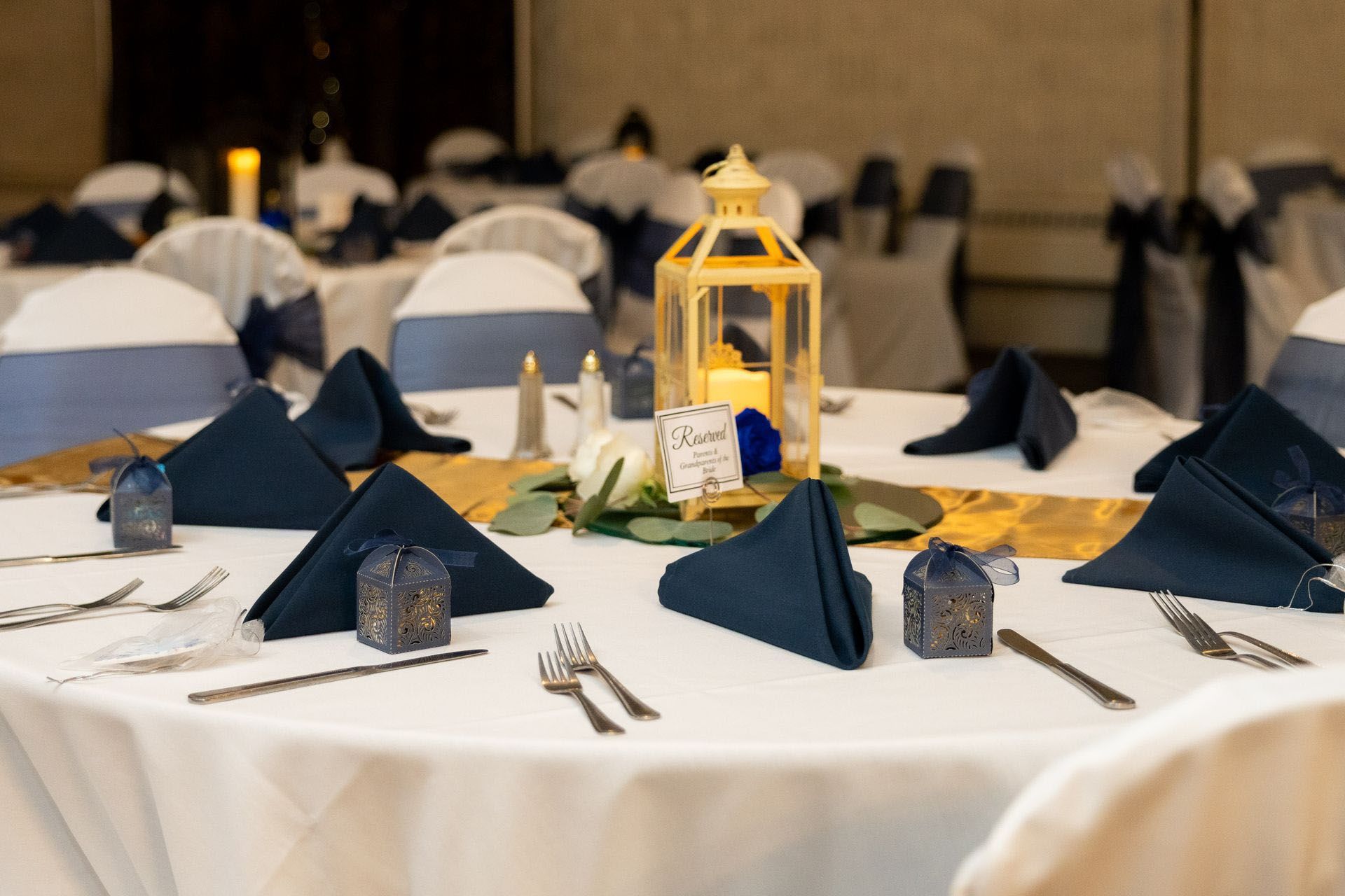 Round banquet table with navy napkins, white tablecloth, and gold lantern centerpiece in a reception hall