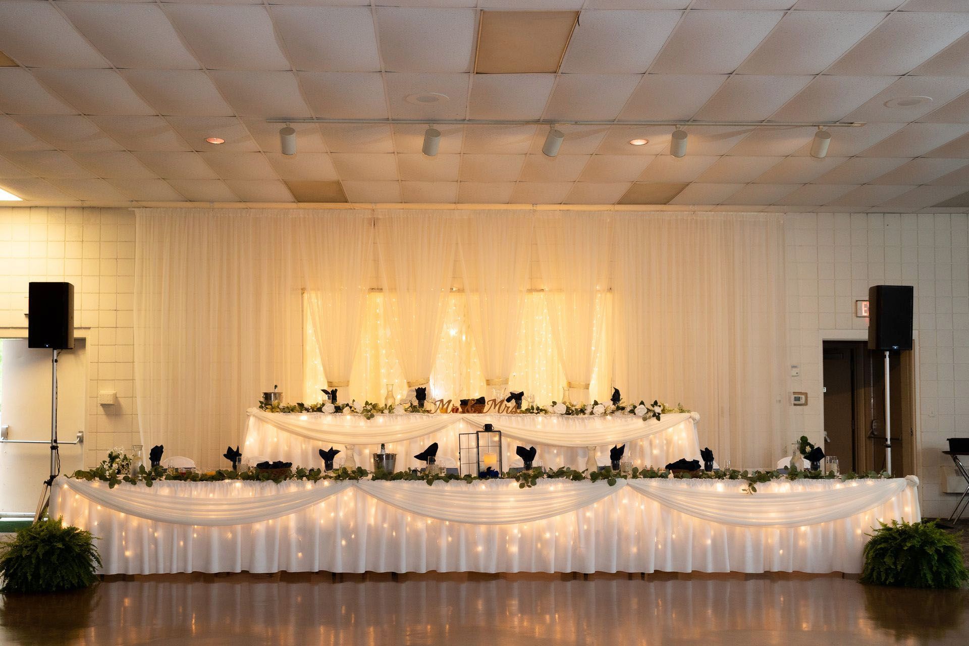 Decorated wedding reception stage with white draping, string lights, and floral arrangements