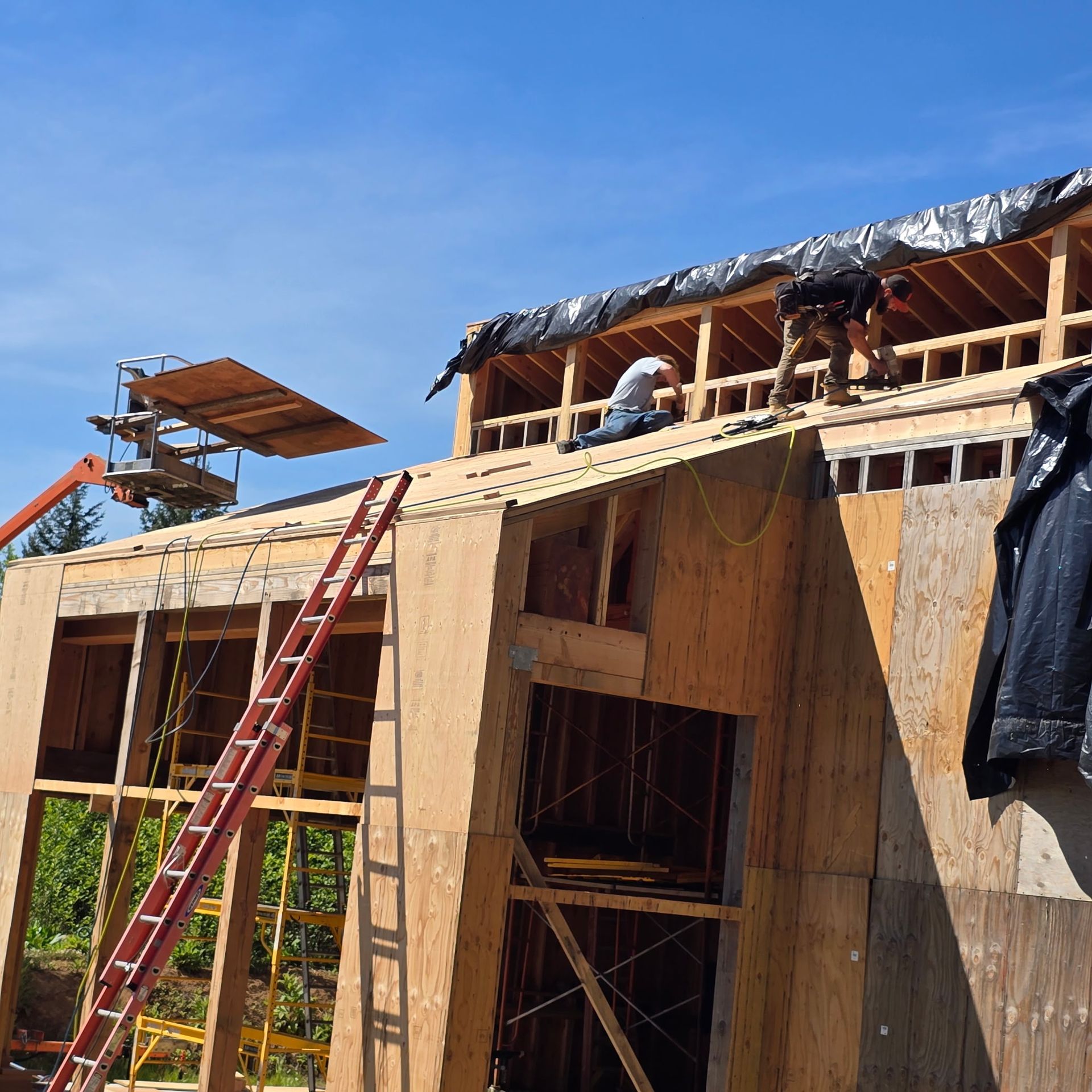 Workers framing a wooden house under construction with ladders and rooftop tarps under a blue sky