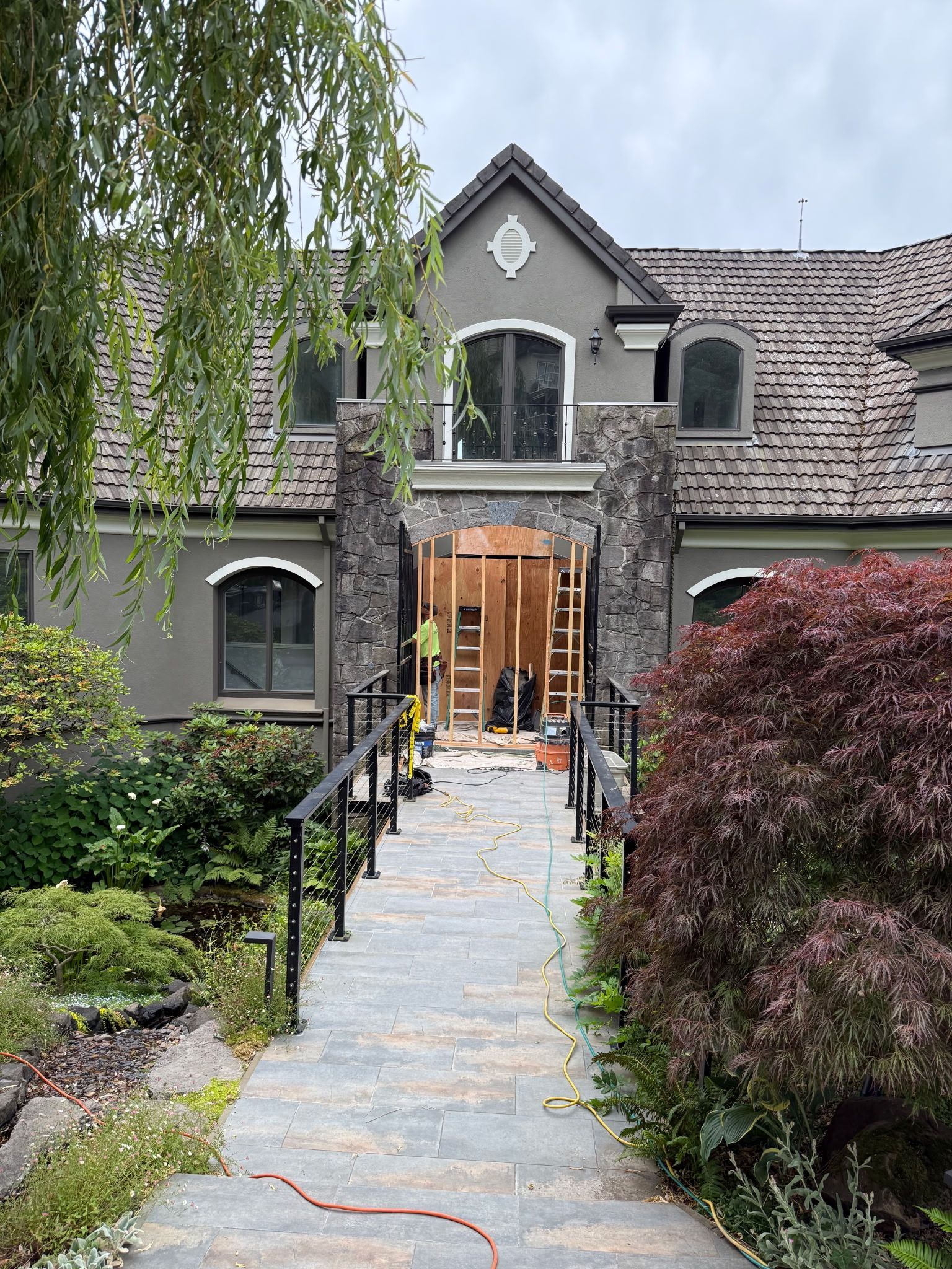 Stone walkway to a gray house with lit wooden front doors, framed by greenery and black railings.