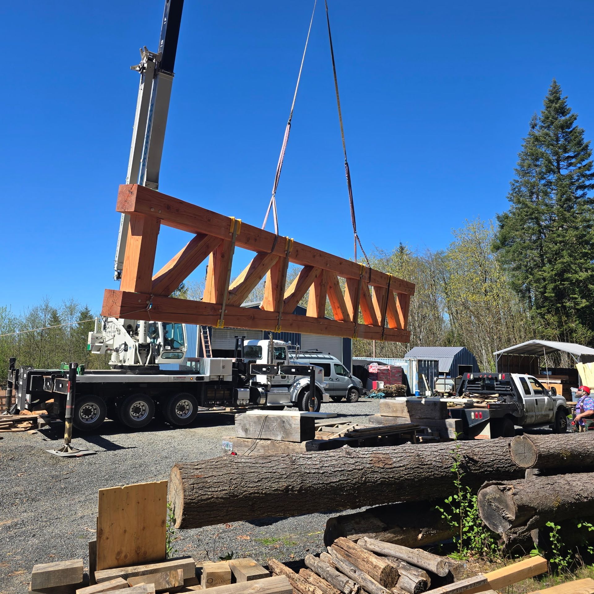 A crane lifts a large wooden timber truss against a clear blue sky at a construction site with logs and work trucks nearby.