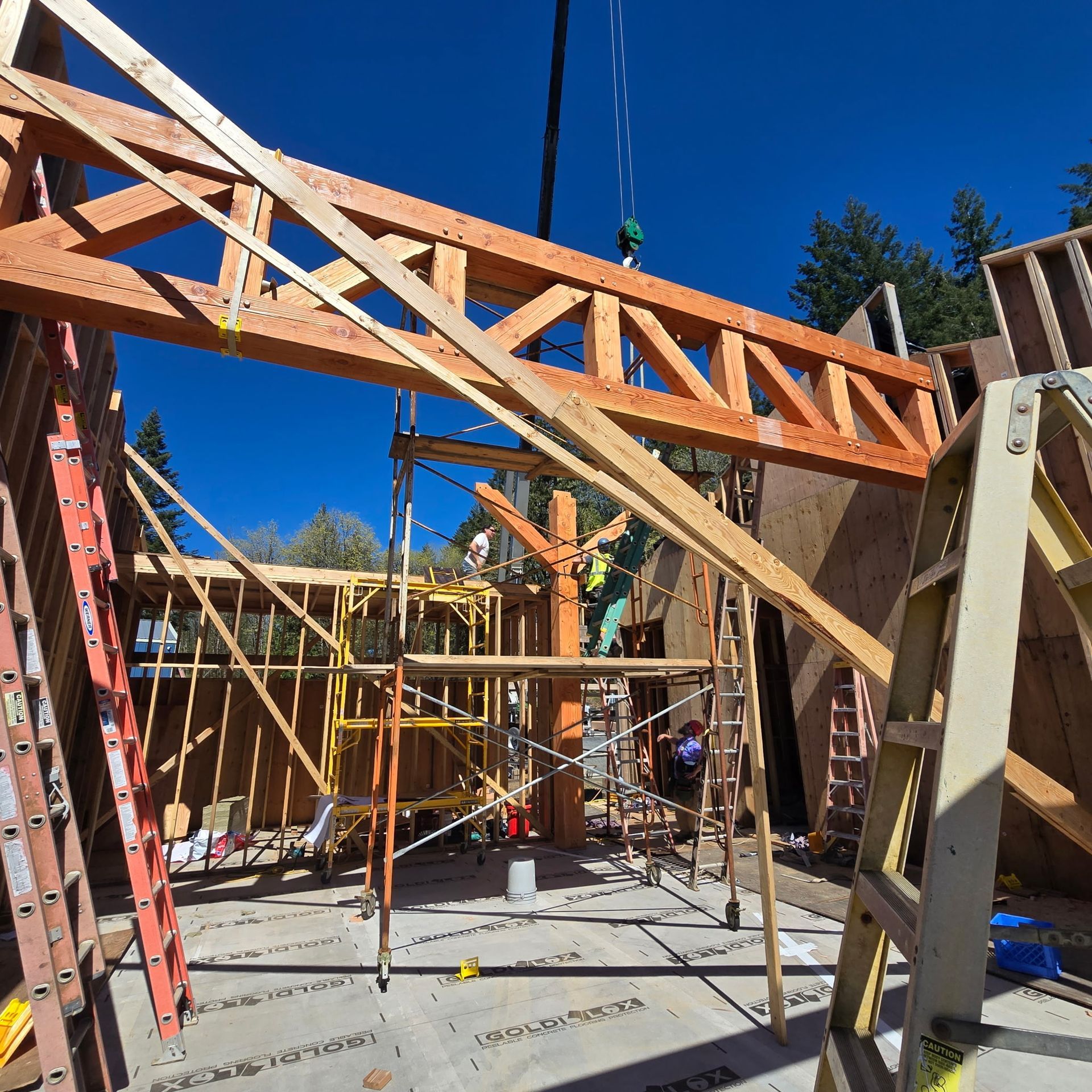 A crane lifts a wooden roof truss into position over a construction site with scaffolding and building frames.