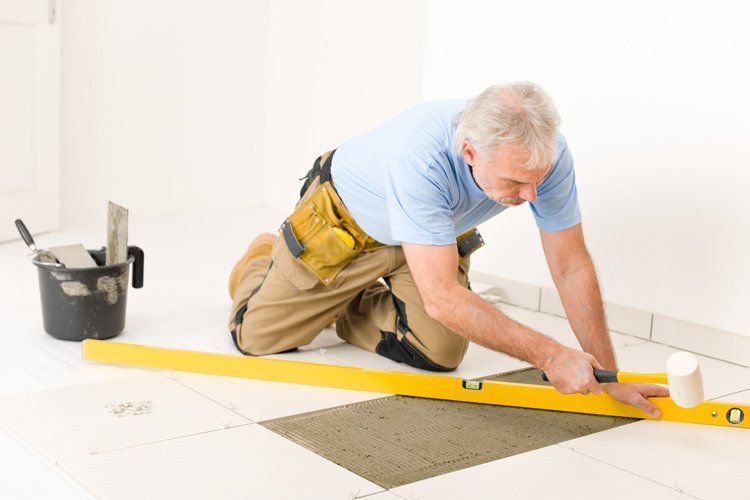 A person kneeling, leveling tile with a yellow level and mallet in a room with white walls.
