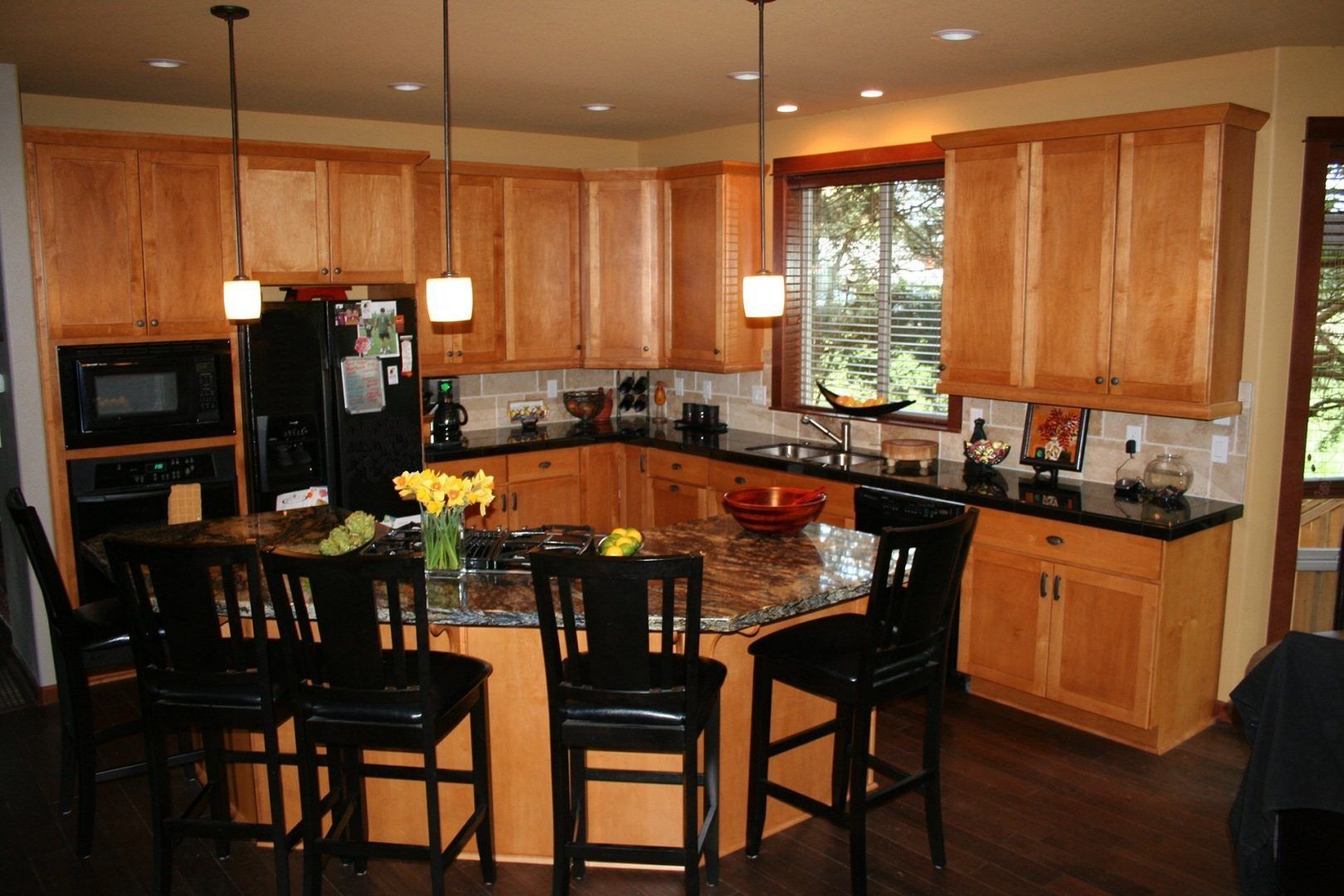 Kitchen with light wood cabinets, black countertops, island with seating, and dark floors.