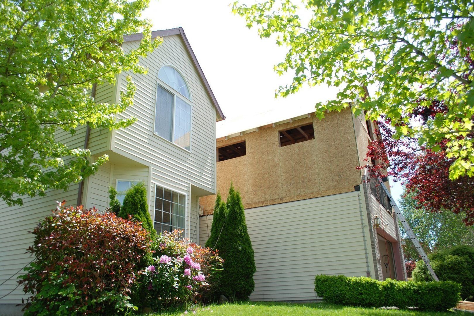 Two-story beige house with a partially constructed addition, surrounded by green trees and bushes.
