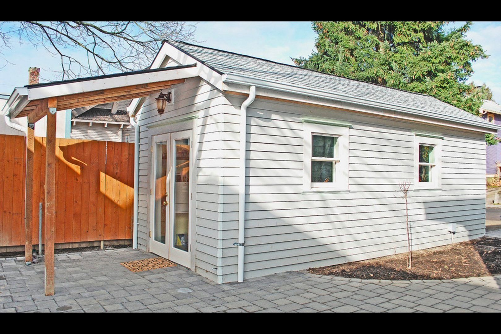Small, light-gray shed with gable roof, French doors, and two windows. Wooden pergola on left side, brick patio.