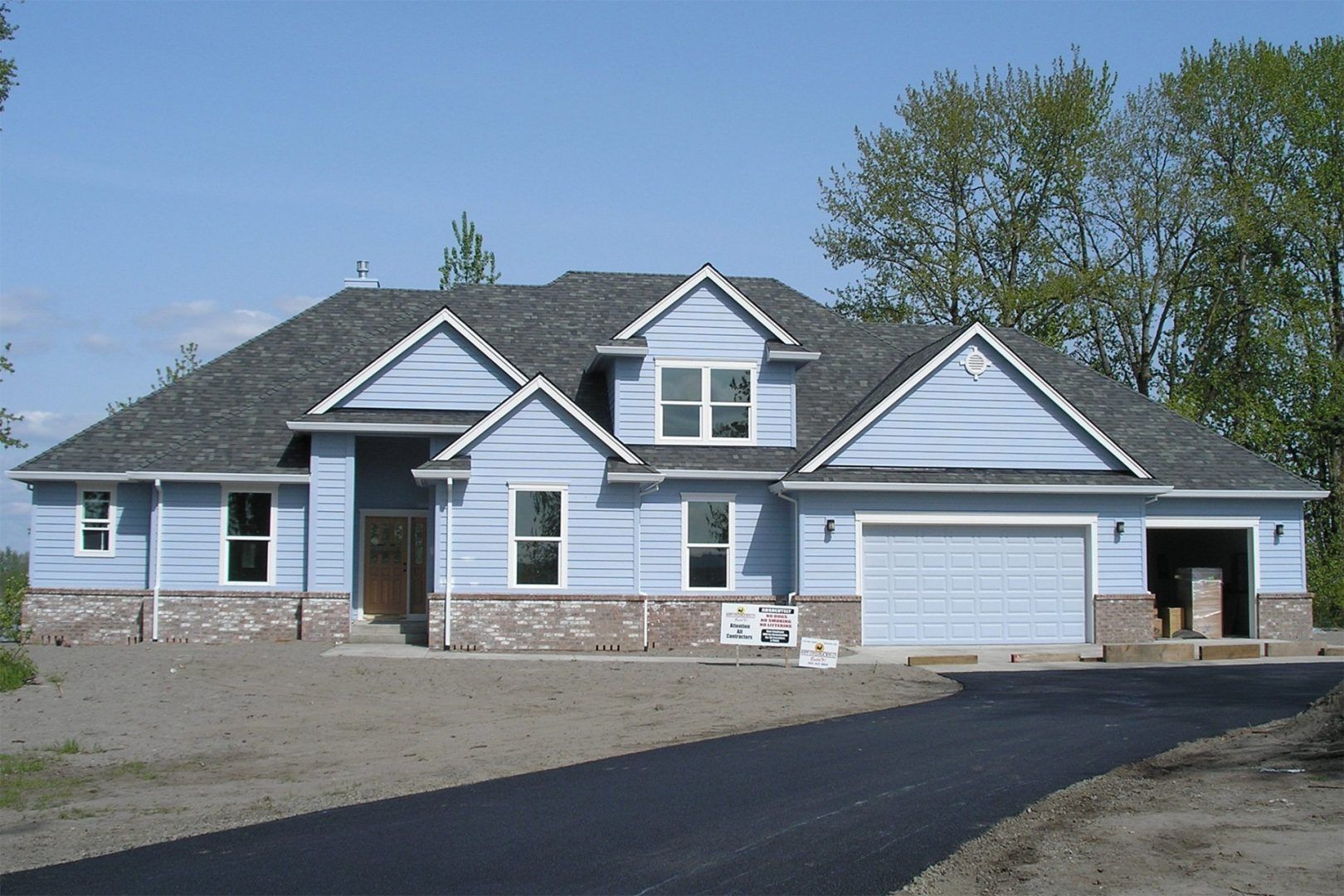 Blue house with grey roof and brick accents, asphalt driveway, unfinished landscaping.