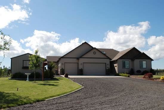 Tan house with a two-car garage, driveway, and green lawn under a partly cloudy blue sky.