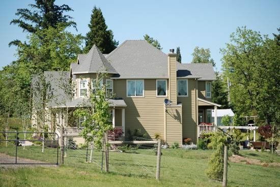 Beige two-story house with gray roof, surrounded by green trees and grass on a sunny day.