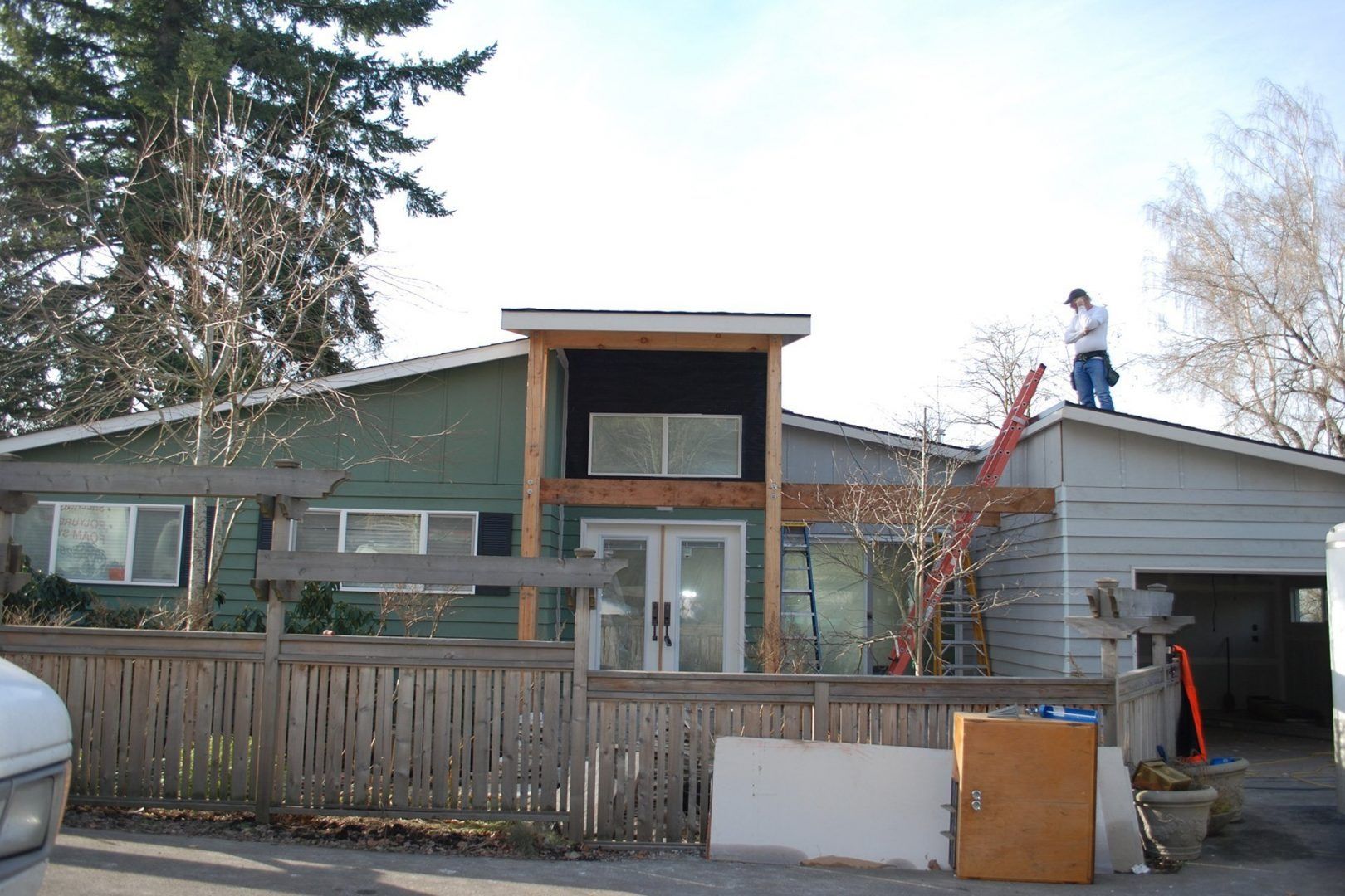House exterior with a person on the roof. Green and gray siding, wooden fence, cloudy sky.