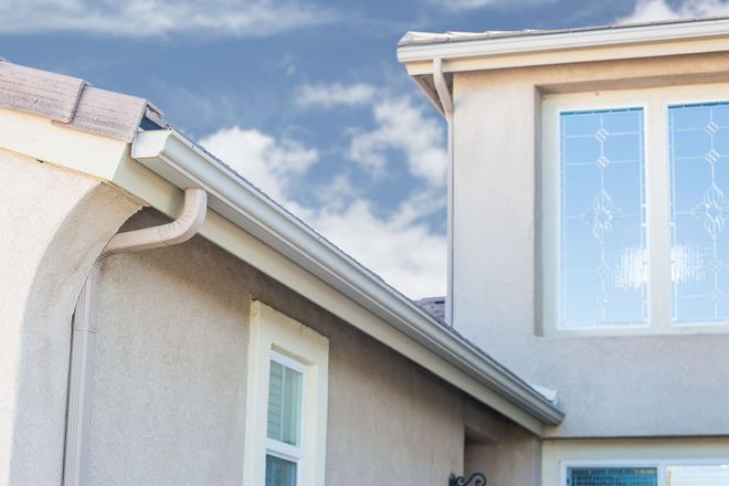 Exterior view of a beige house with a gutter, white-framed windows, and a blue cloudy sky.