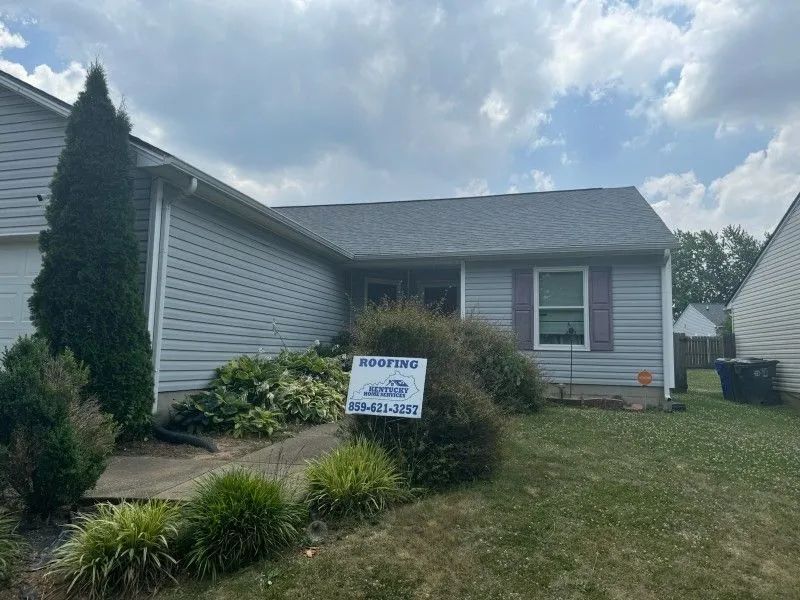 Gray house with new roof; sign in front, cloudy sky.