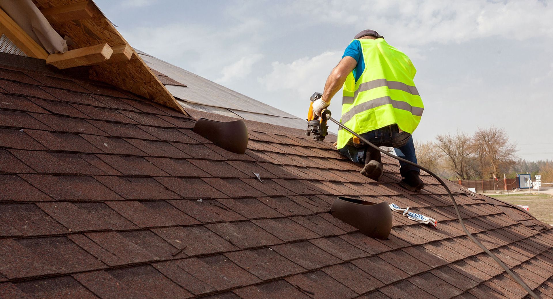 Roofer in yellow vest using a nail gun on a brown shingle roof under a cloudy sky.