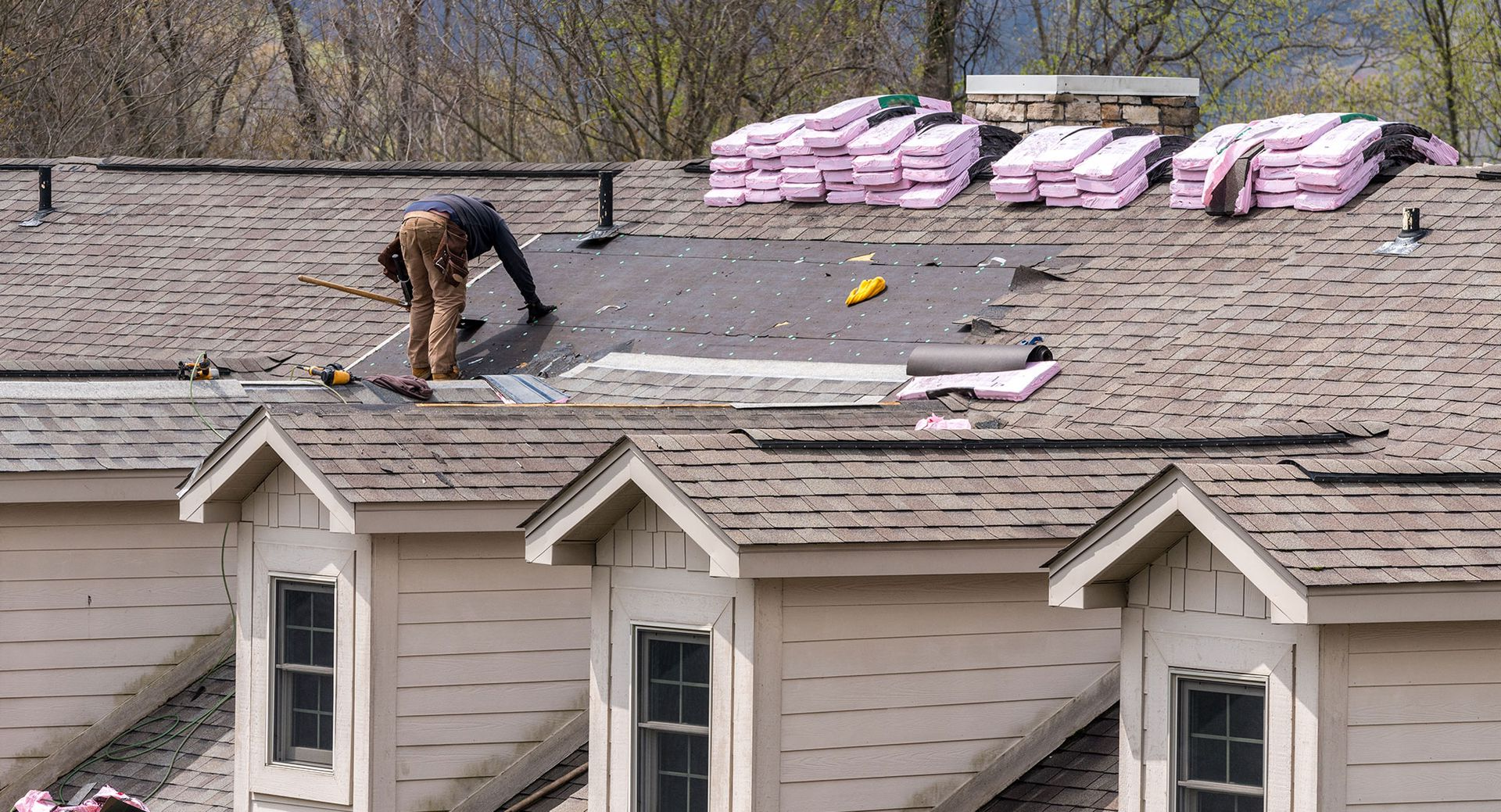 Roofer replacing shingles on a house roof. Pink insulation and supplies are visible nearby.
